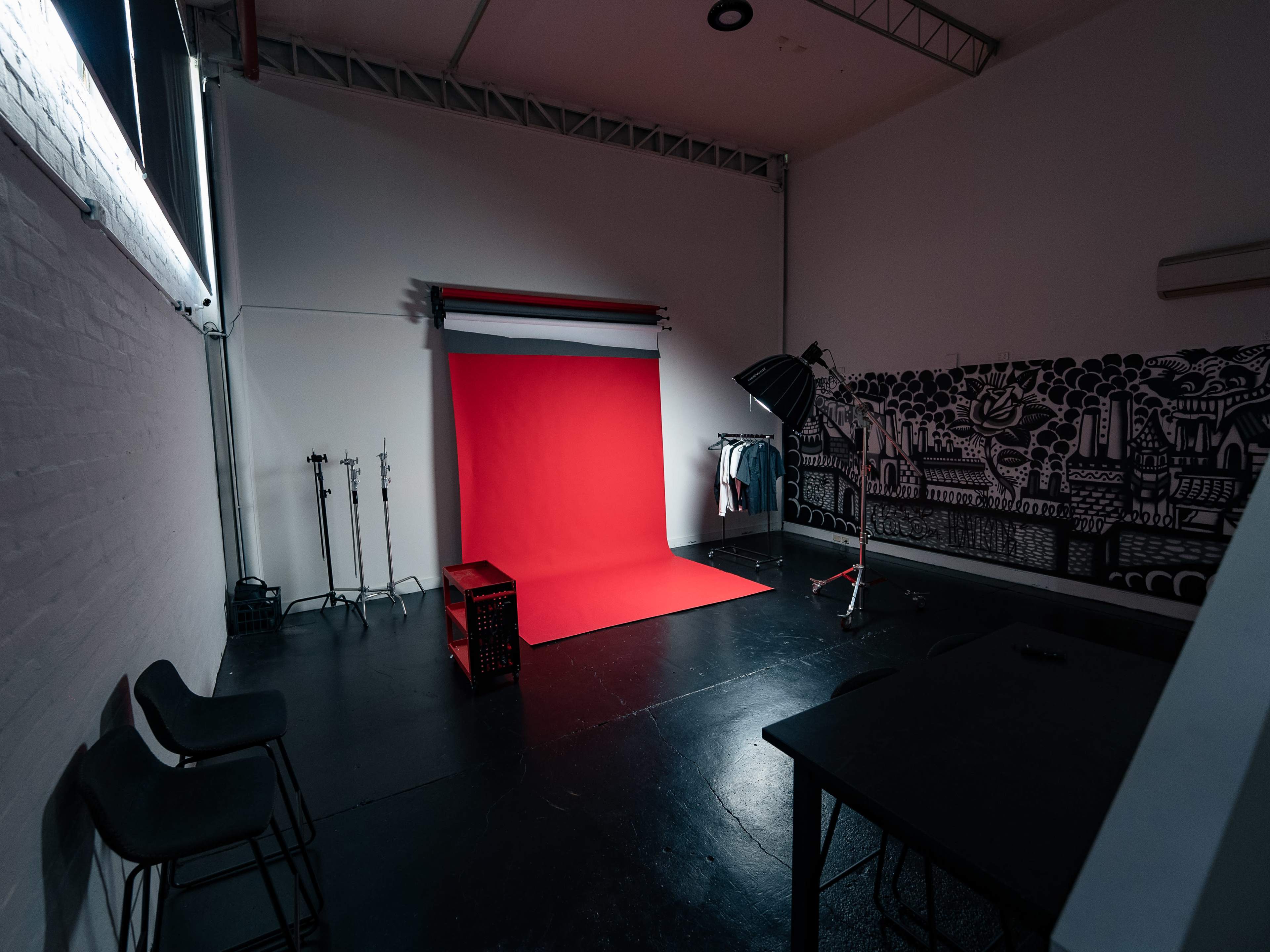 The image shows a photography studio with a red backdrop, a clothing rack, a light stand, and dark flooring.