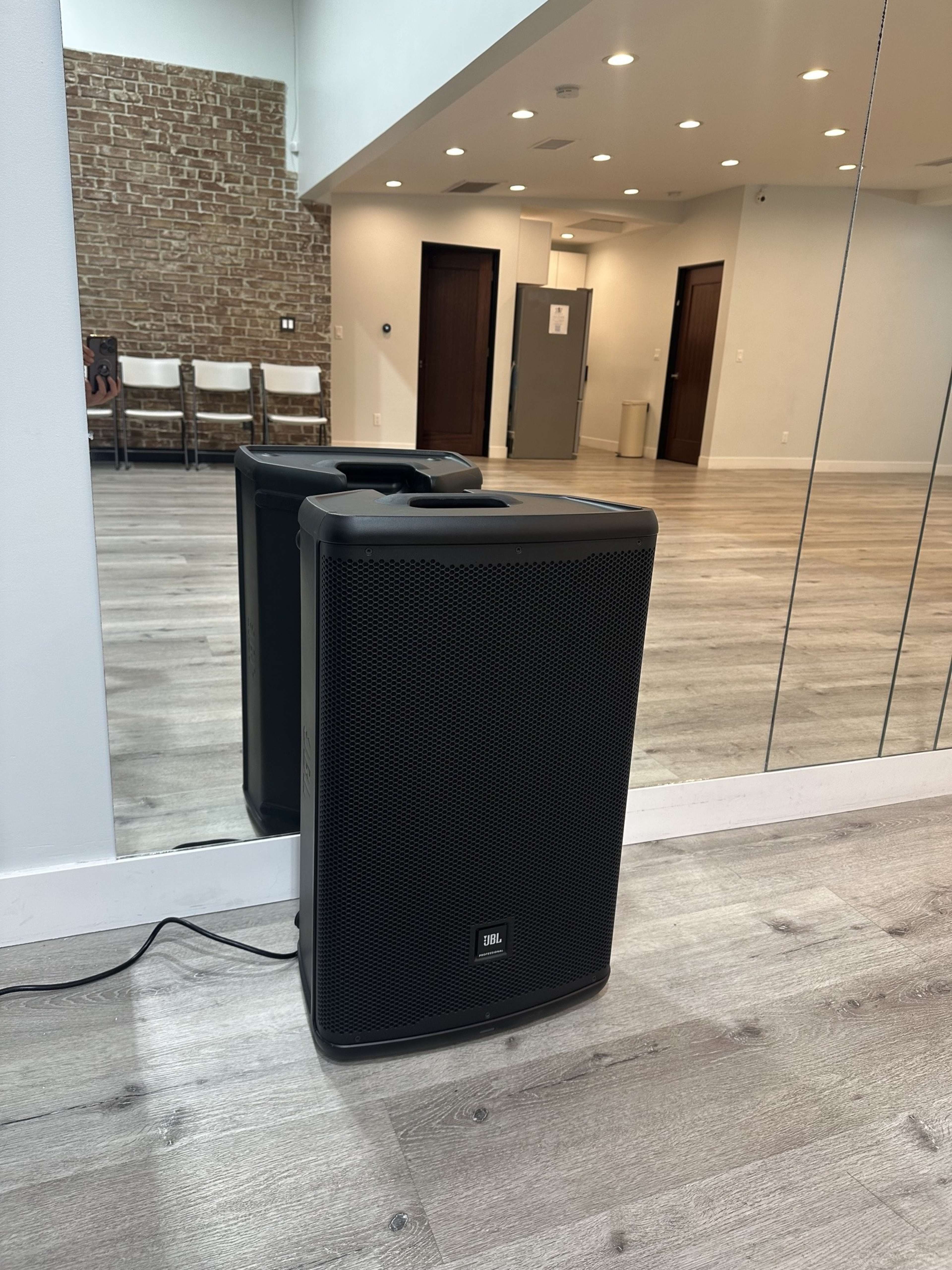 A black JBL speaker is positioned on the floor in a well-lit room with wooden flooring and a mirrored wall.