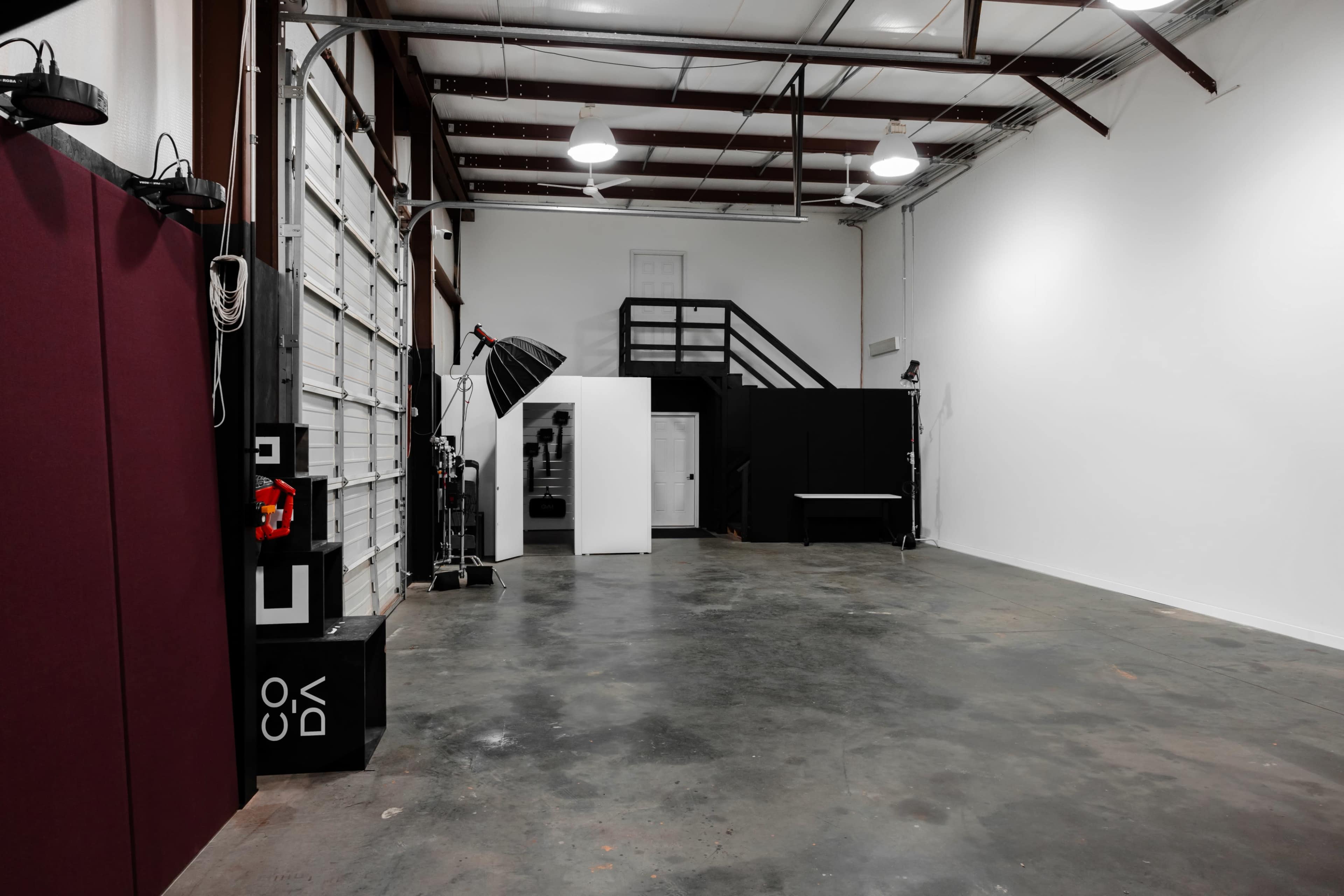 A spacious, empty studio with a concrete floor, white walls, and various lighting equipment arranged along one side.