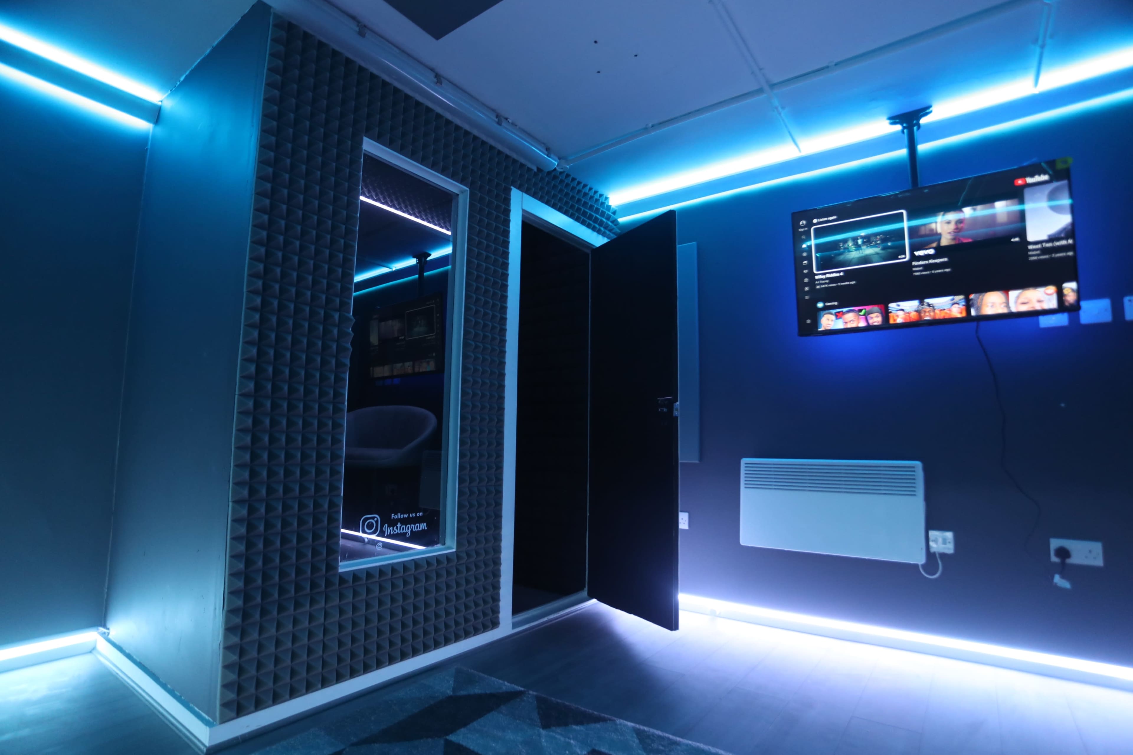 A small room with soundproofed walls, a door slightly ajar, and a television mounted on the wall displaying content, illuminated by blue LED lights.