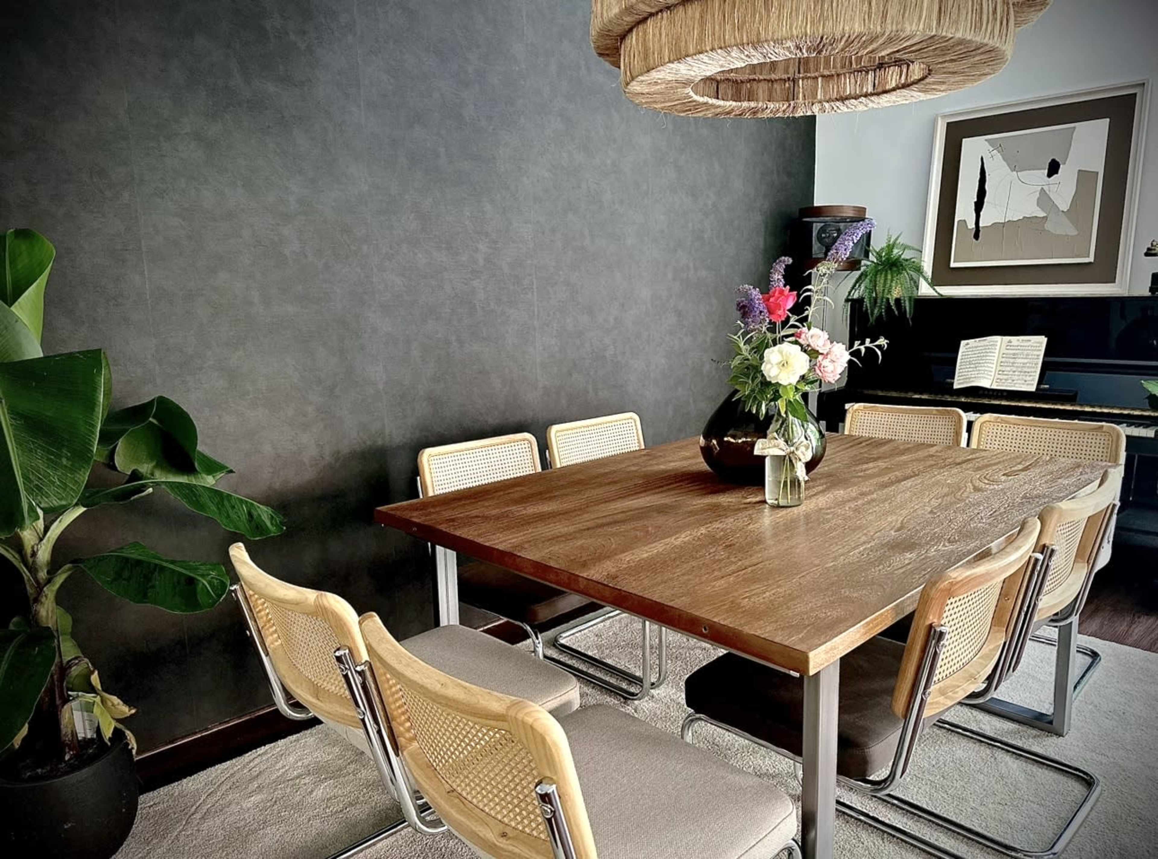 A wooden dining table surrounded by eight chairs is set in a room with a dark wall, a large plant, and a hanging light fixture.