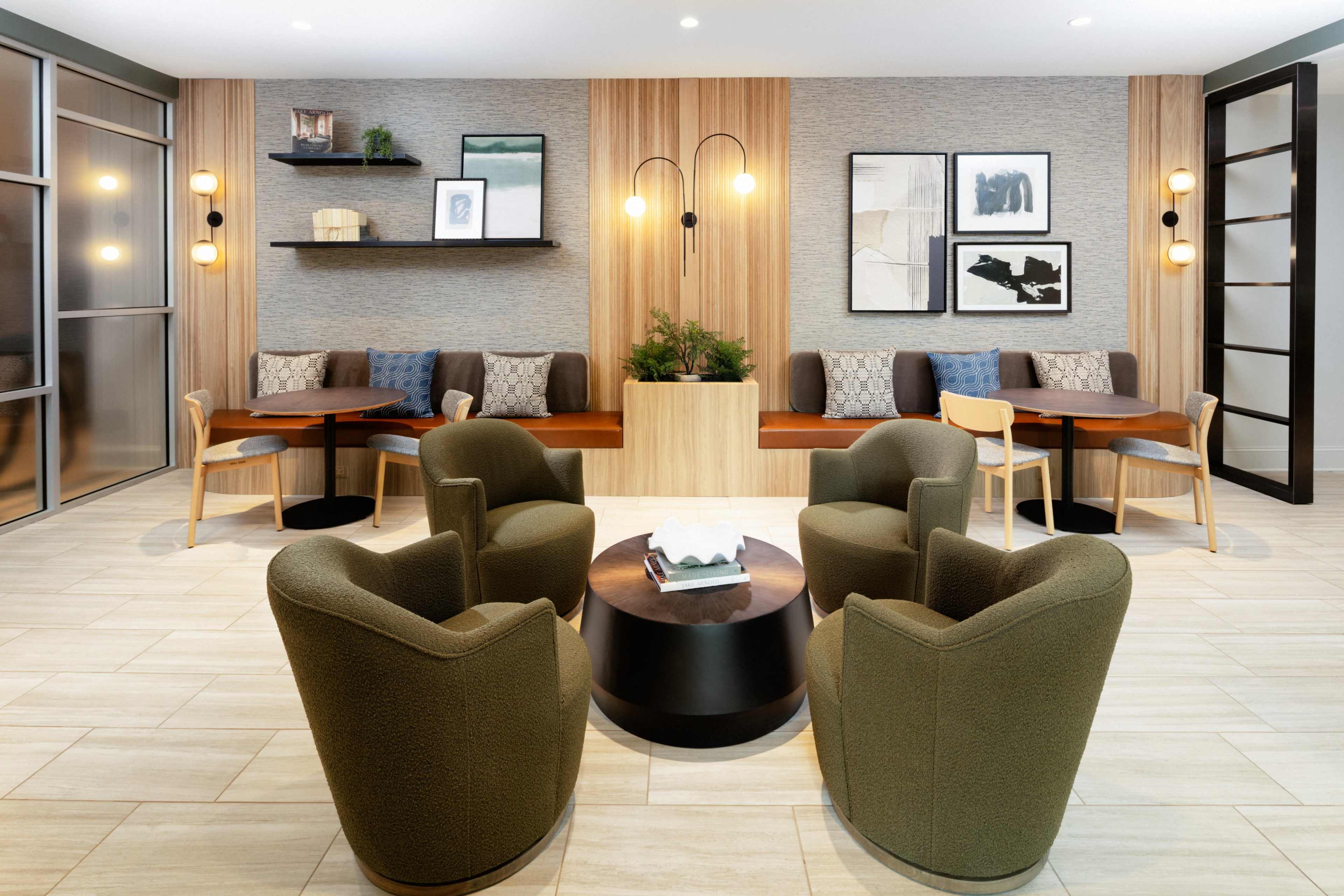 The image shows a modern lounge area featuring two circular tables surrounded by four green armchairs, with a wooden accent wall and framed artwork in the background.