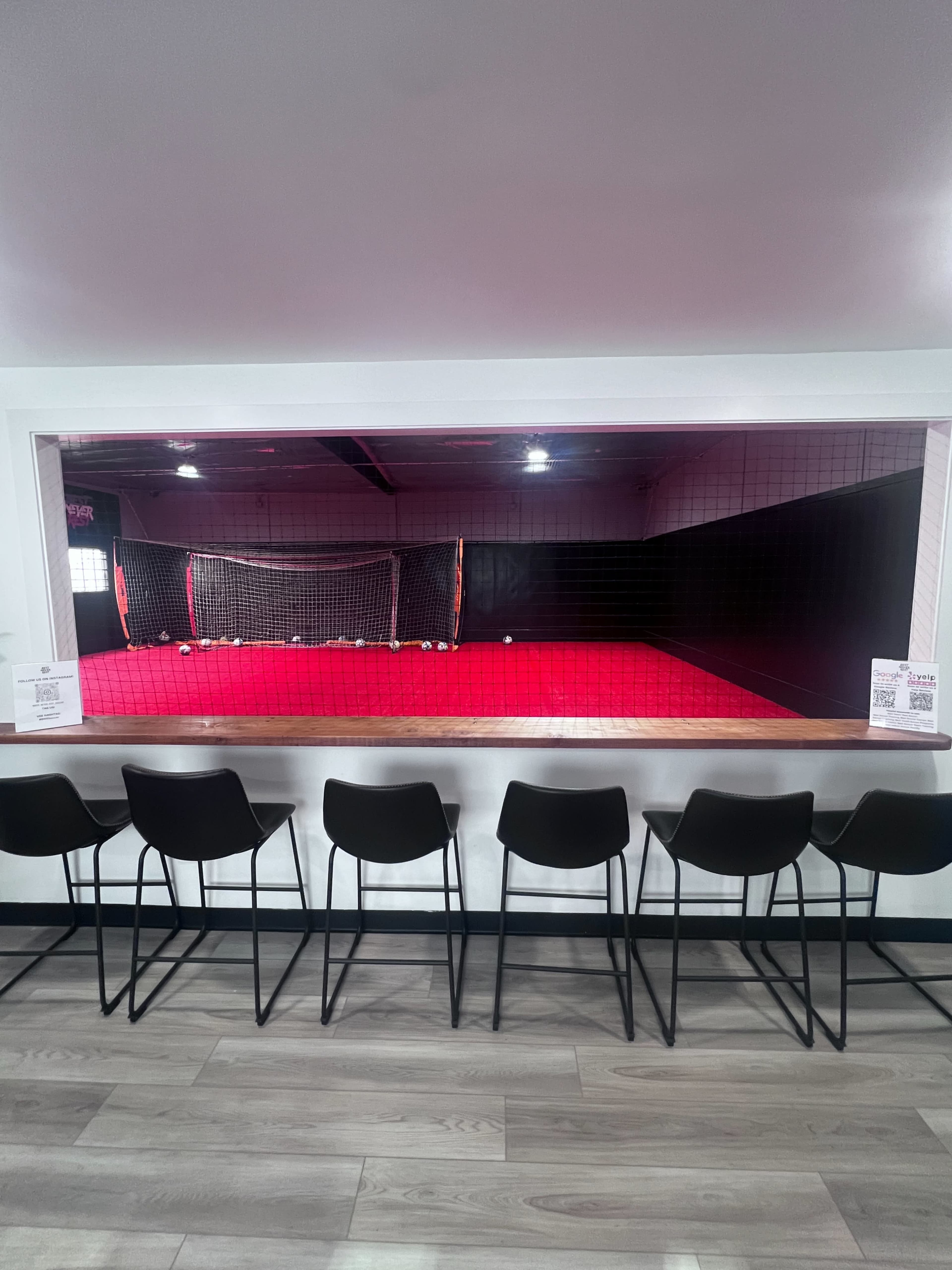 The image shows a modern facility with a row of black chairs facing an indoor soccer field, separated by a glass window.