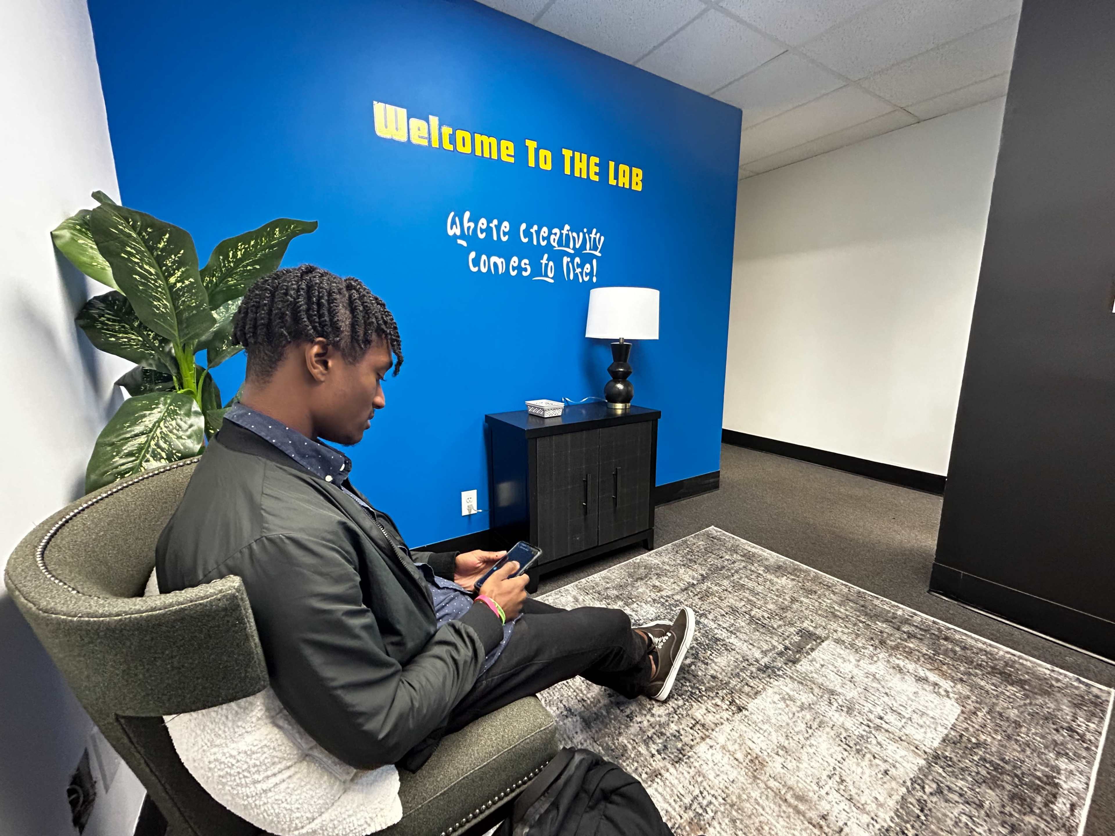 A young man sits on a chair while using his phone in a room with a blue wall that features the phrase "Welcome To THE LAB" and a bedside lamp.