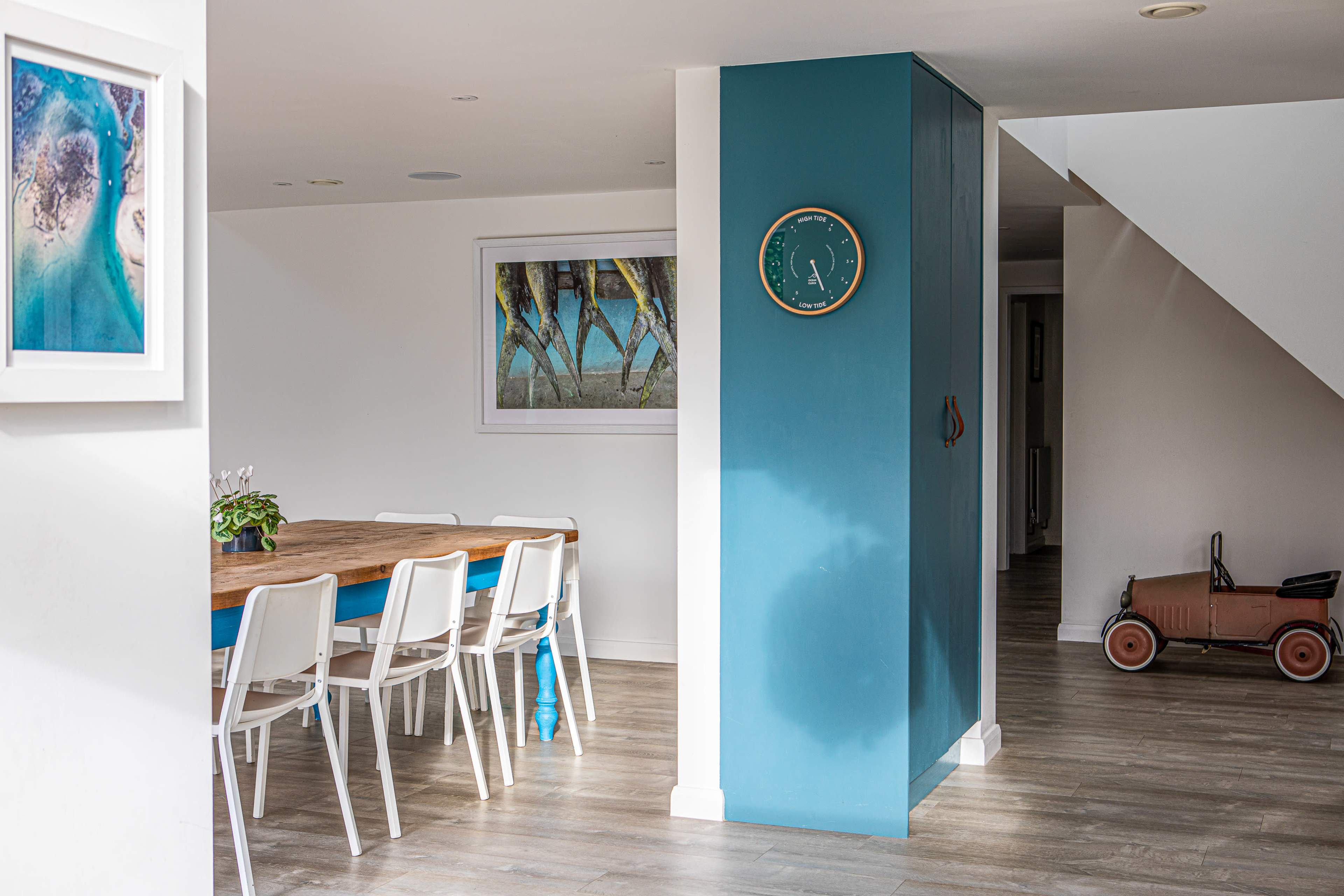 A modern interior featuring a dining area with a wooden table and white chairs, a blue accent wall with a clock, and a vintage toy car in a hallway.