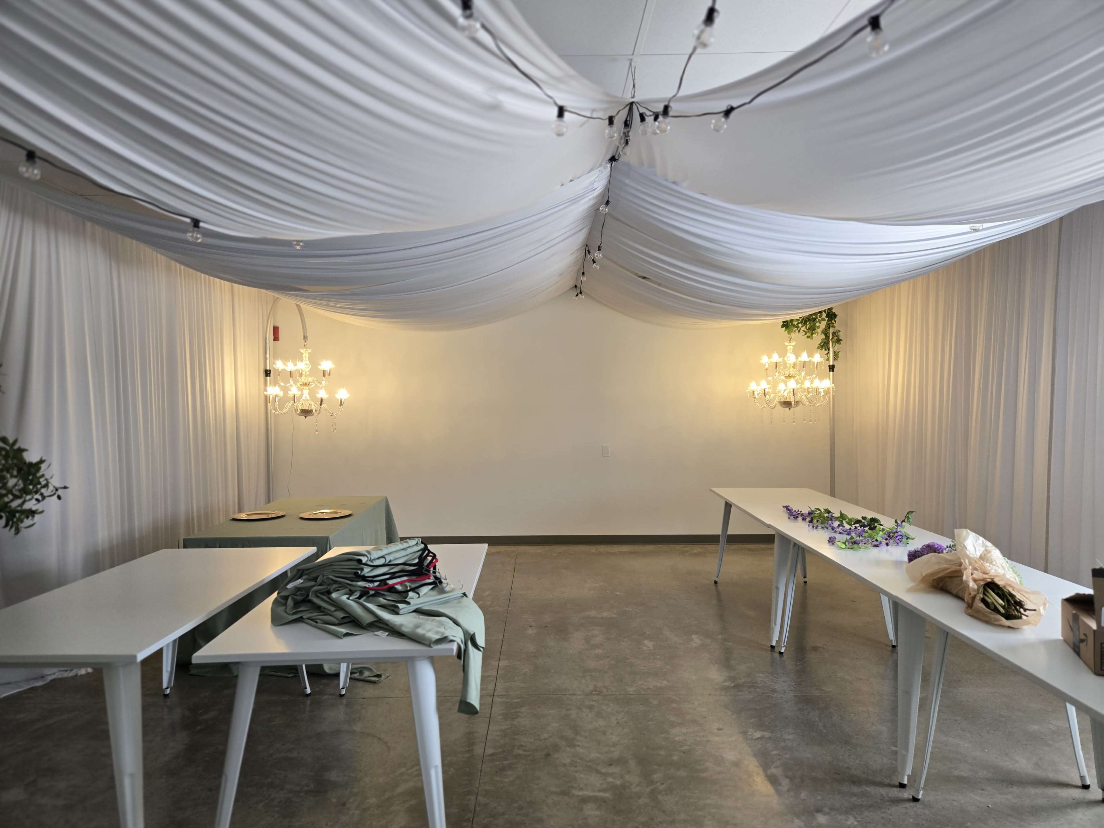A spacious interior features draped white fabric overhead, two chandeliers on the walls, and several tables with various items arranged on them.