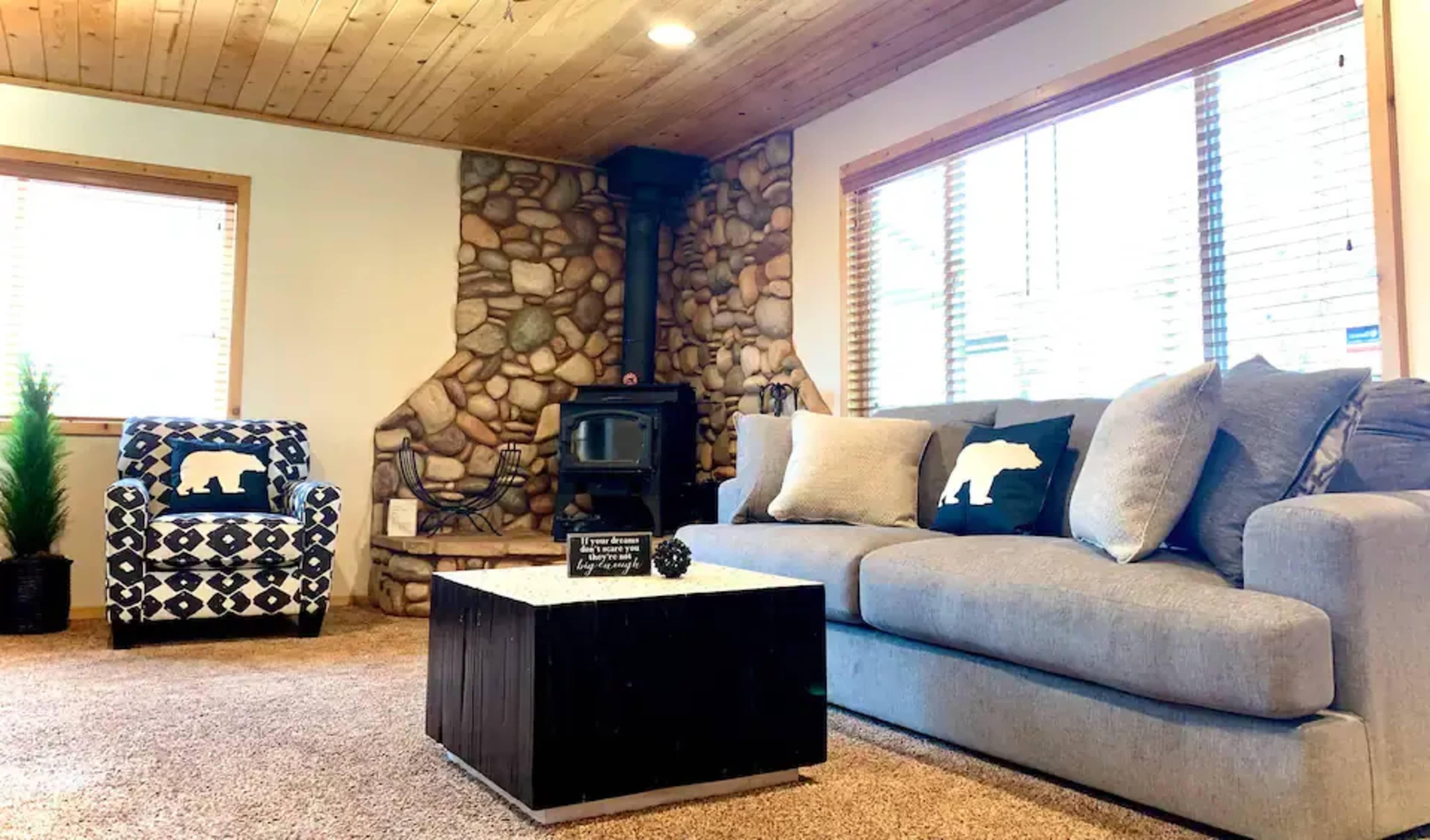 The living room features a gray sofa, a black and white patterned armchair, a wooden coffee table, and a stone fireplace with a wood stove.