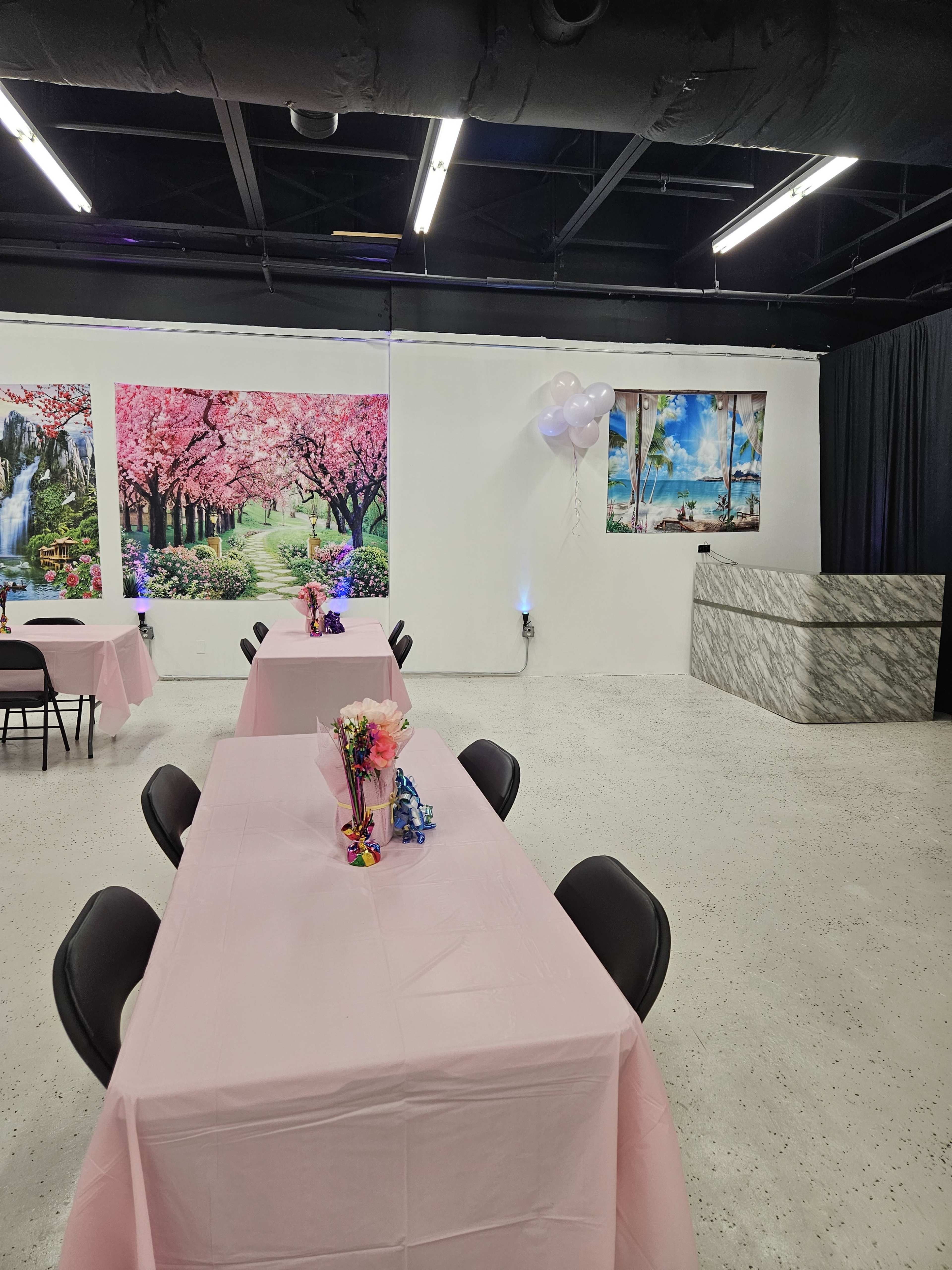 A decorated event space with pink tables and floral centerpieces, featuring scenic wall murals and a reception area at the back.