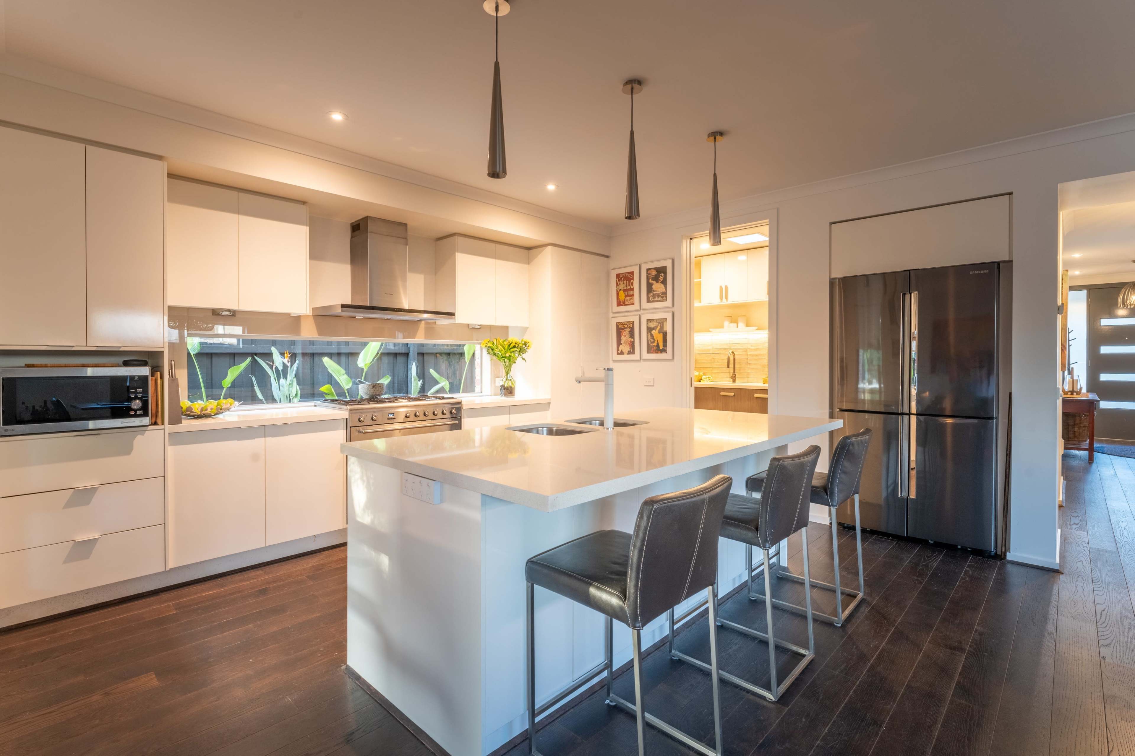 The image shows a modern kitchen with white cabinets, a central island with bar stools, stainless steel appliances, and large windows allowing natural light.