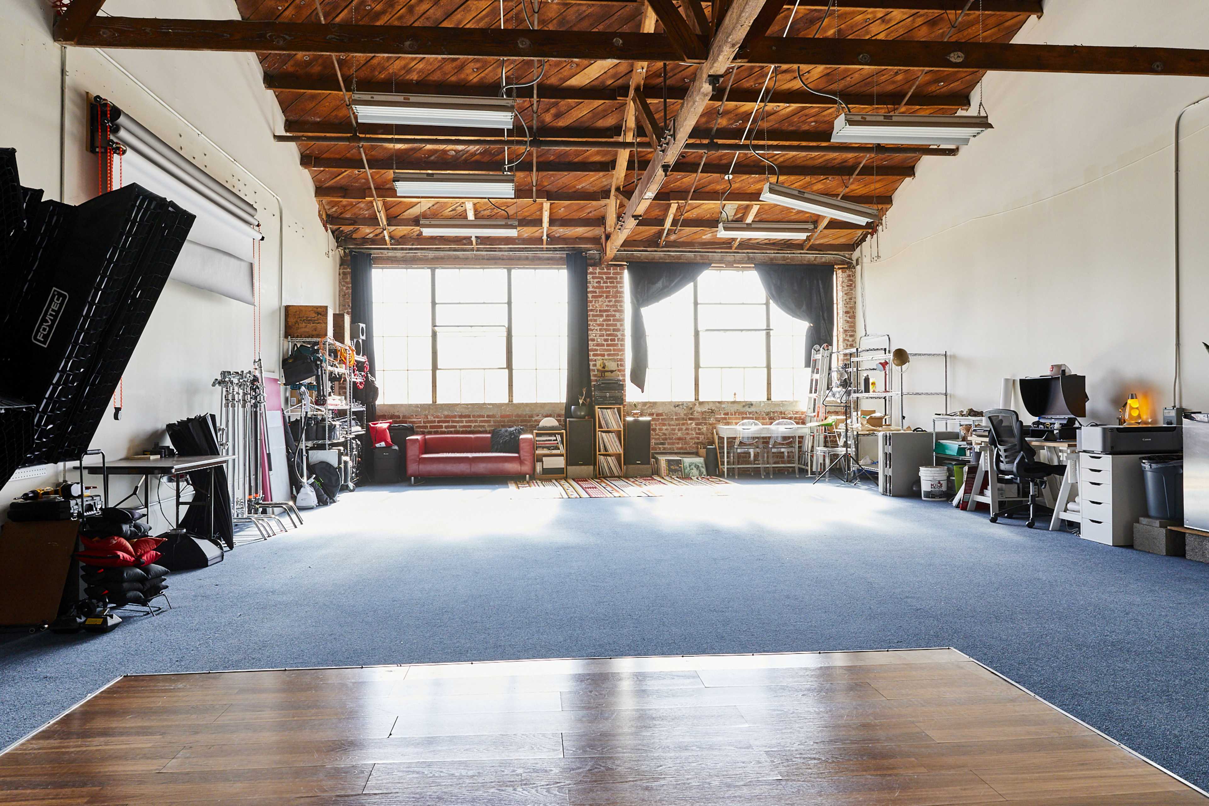 A spacious industrial studio features large windows, exposed brick, wooden beams, and various equipment along the walls.