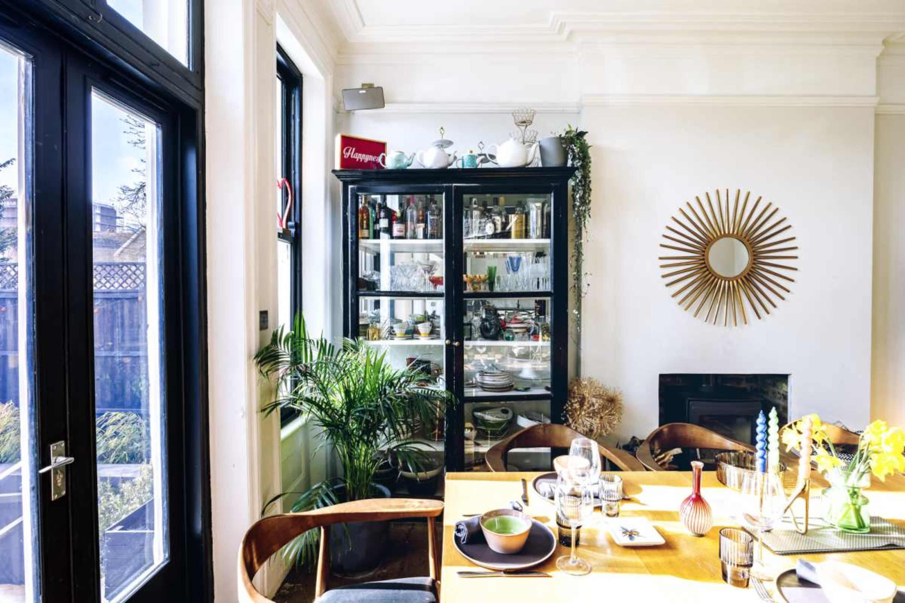 A dining area features a table set with dishes and glasses, alongside a glass-fronted cabinet displaying various items and plants.