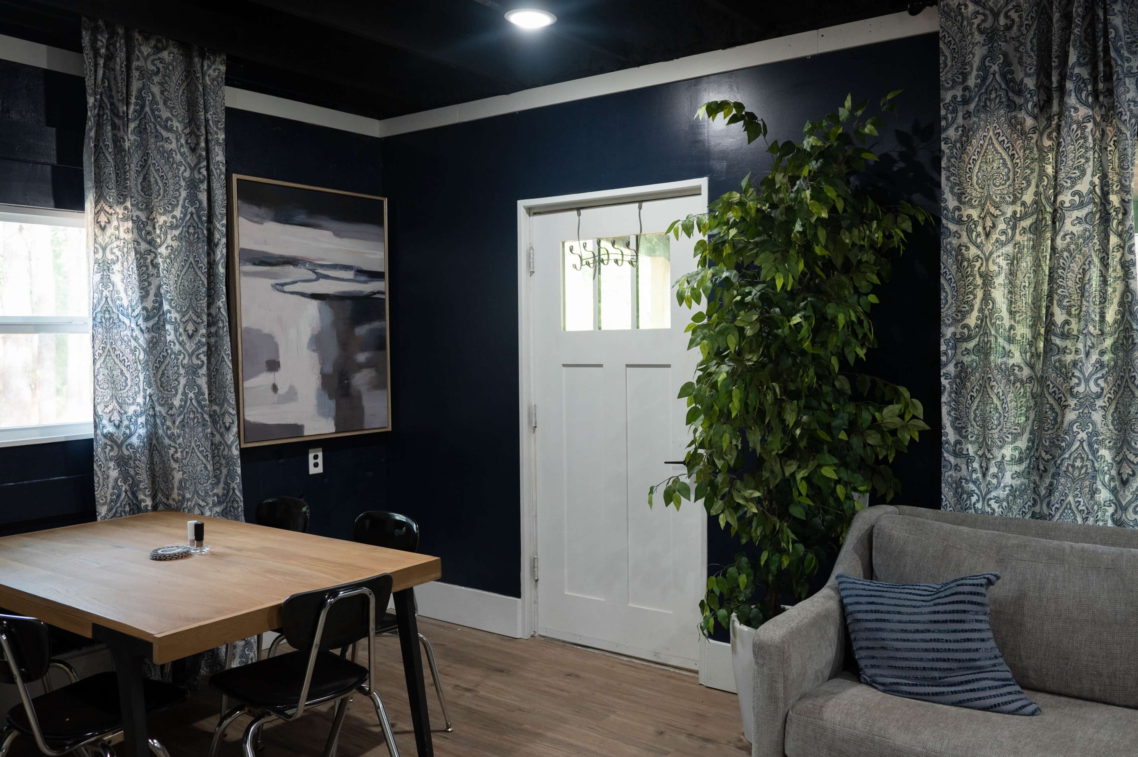 The image shows a living space with a wooden table, a sofa, a potted plant, and patterned curtains, featuring a dark blue wall with framed artwork and a white door.