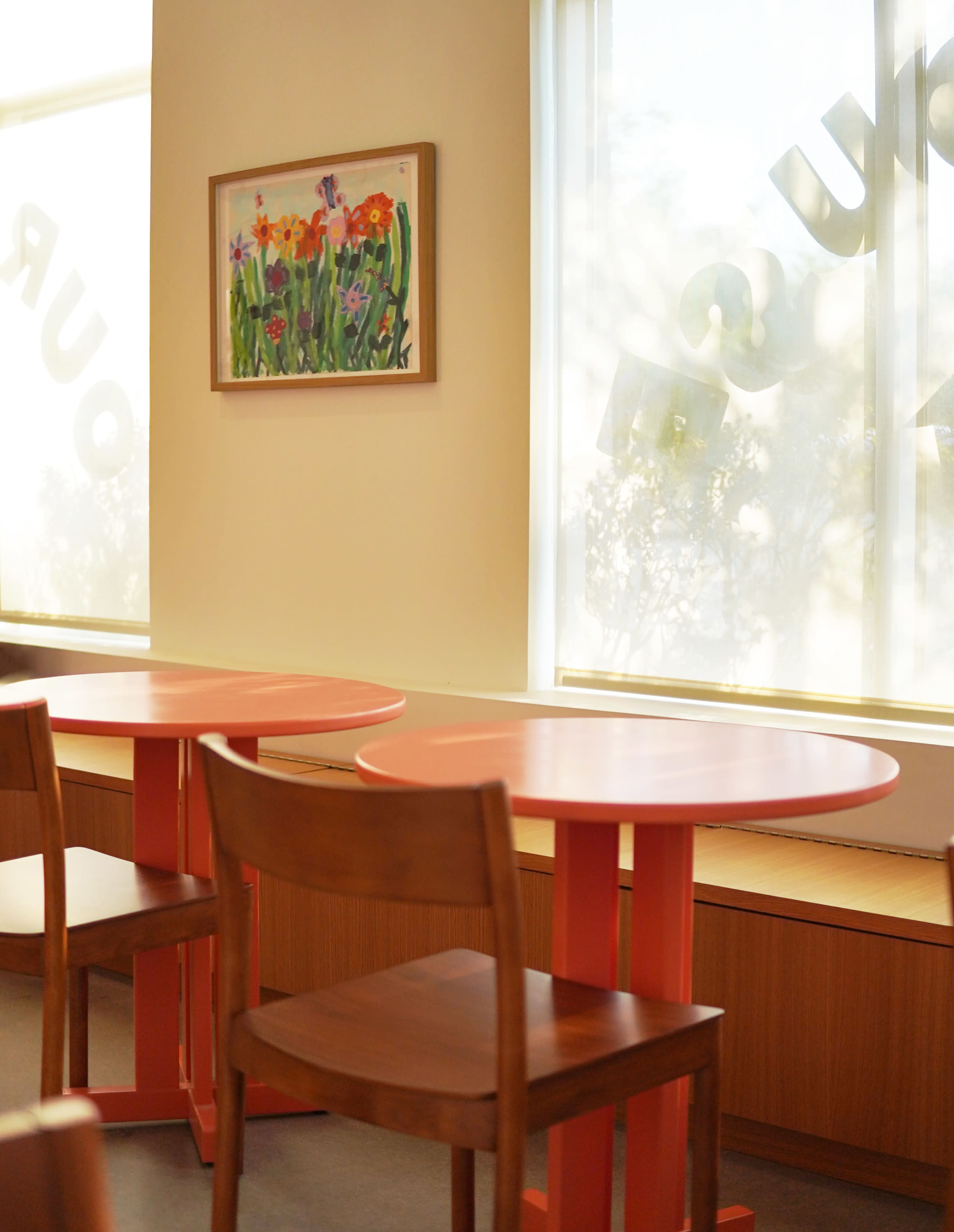 The scene features two round orange tables and wooden chairs in a brightly lit café, with a floral painting hanging on the wall near a large window.