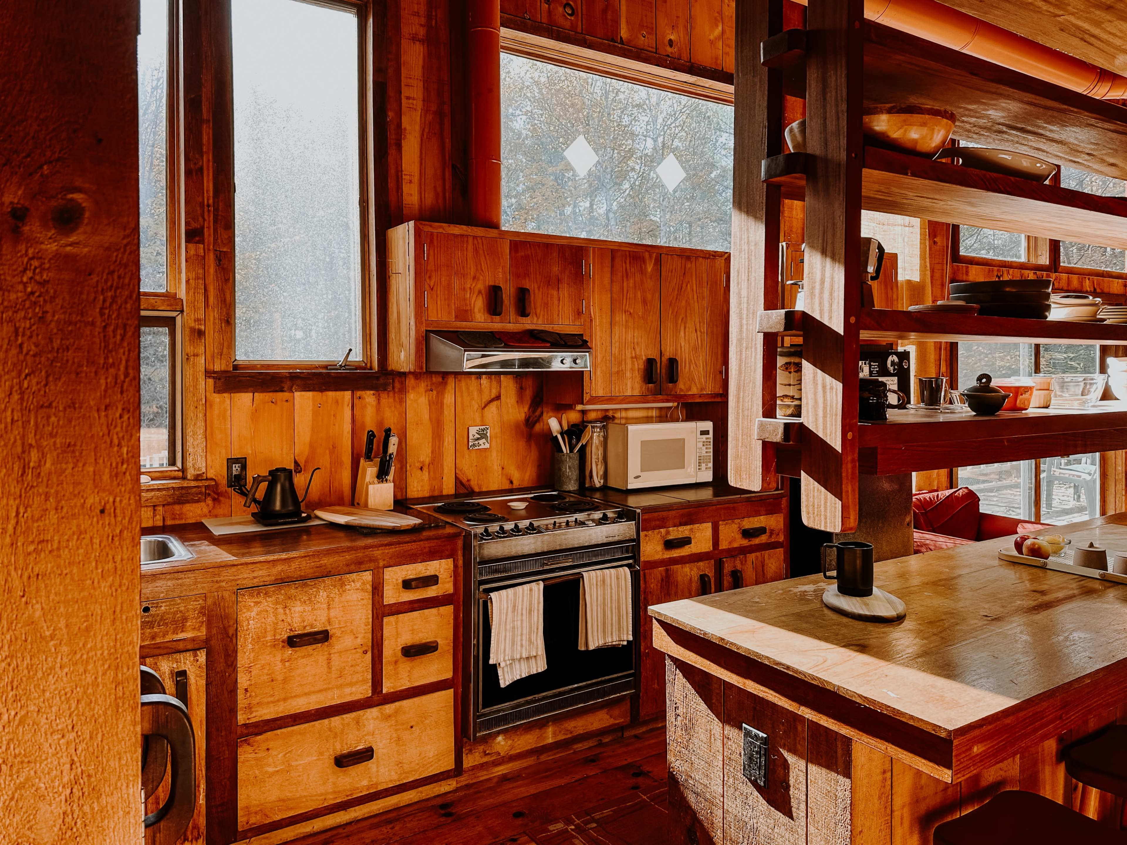 The image shows a rustic kitchen with wooden cabinetry, a stove, a microwave, and large windows allowing natural light to enter.