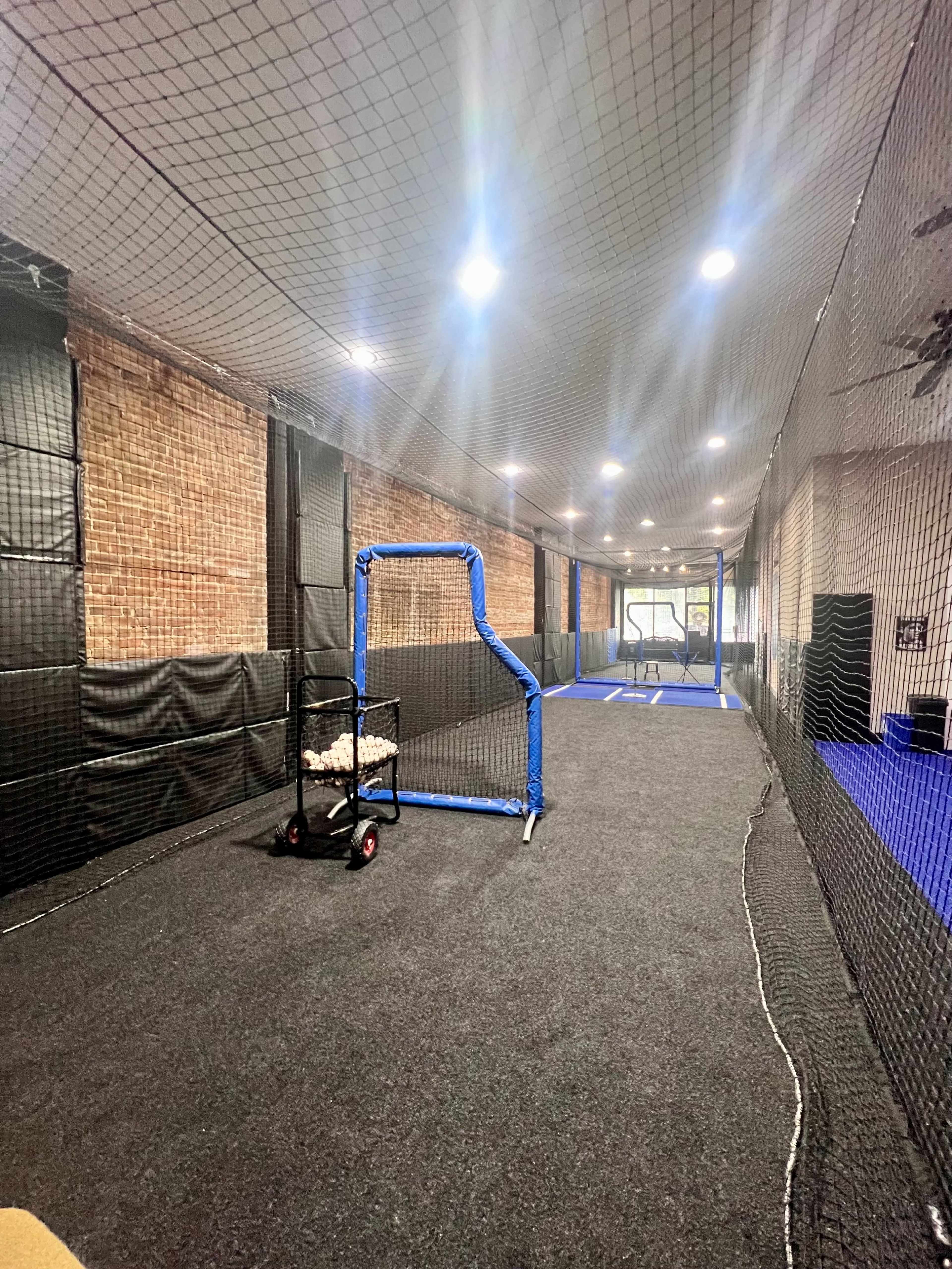 An indoor sports facility with a batting cage, a cart filled with baseballs, and a netted area for practice.