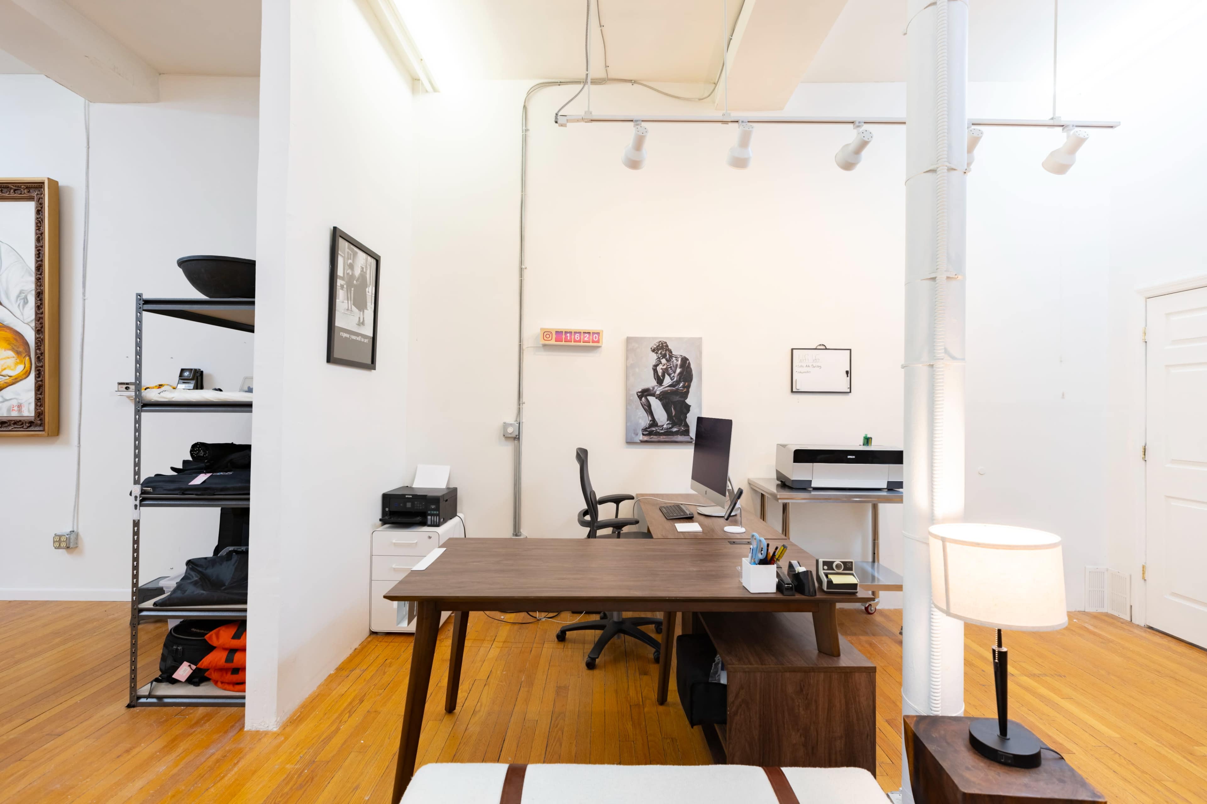 The image shows a bright office space featuring a wooden desk, a printer, shelving with items, and a lamp, all arranged against white walls with hardwood flooring.