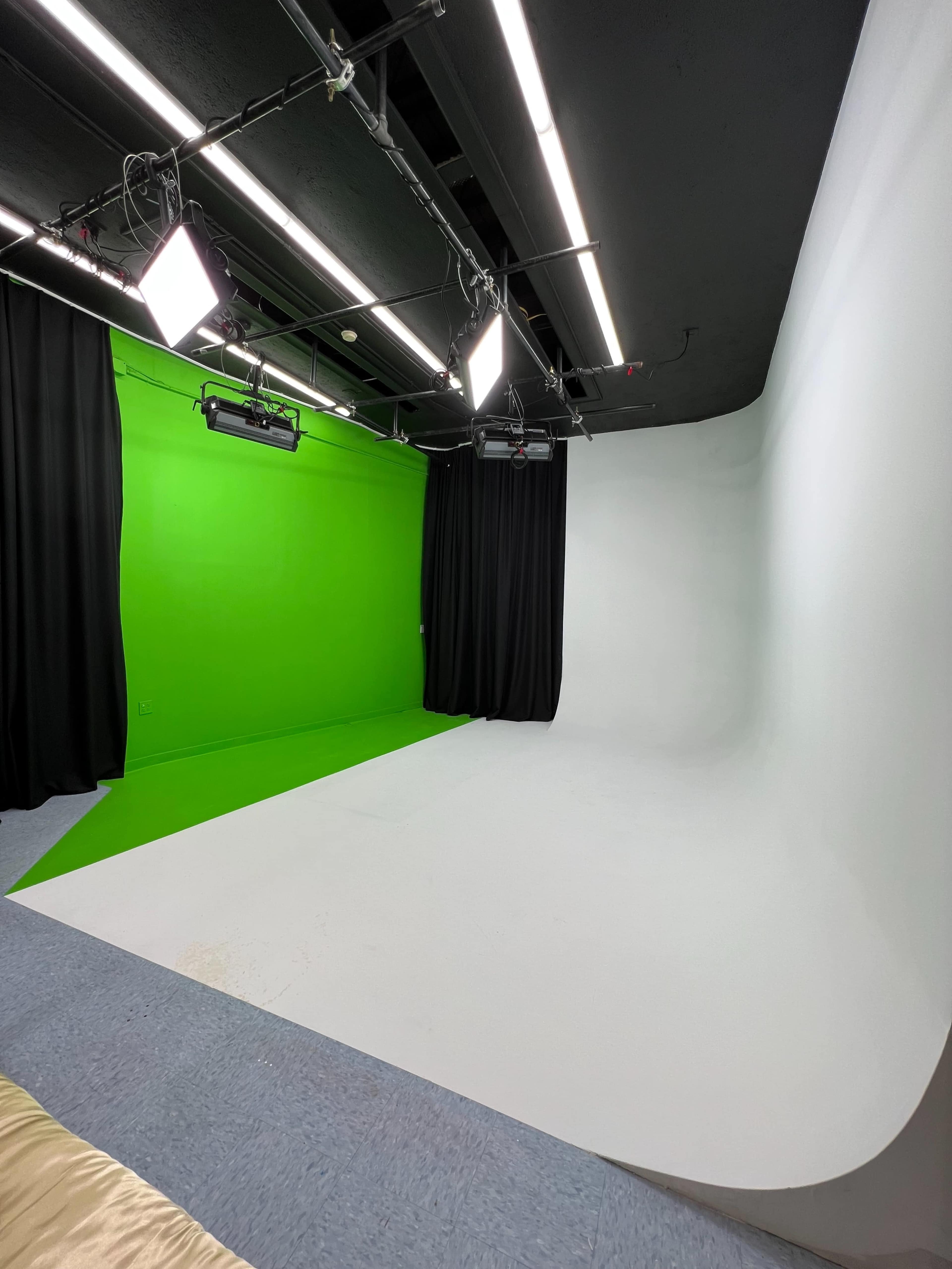 The image shows a photography studio featuring a green screen on one side and a white backdrop on the other, with multiple overhead lights and black curtains.