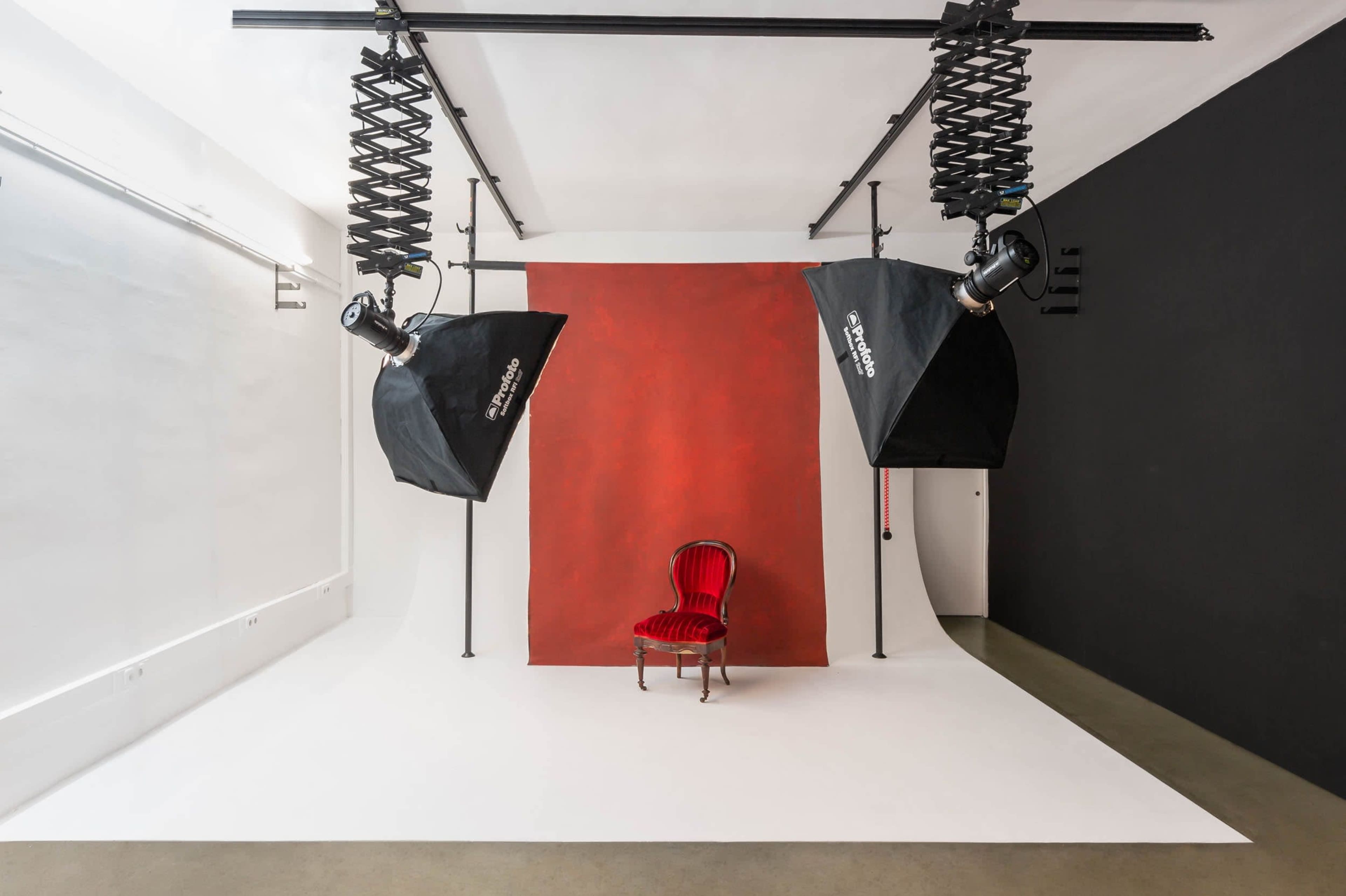 The image shows a photography studio with a red backdrop, two softbox lights, and a single red chair placed on a white floor.