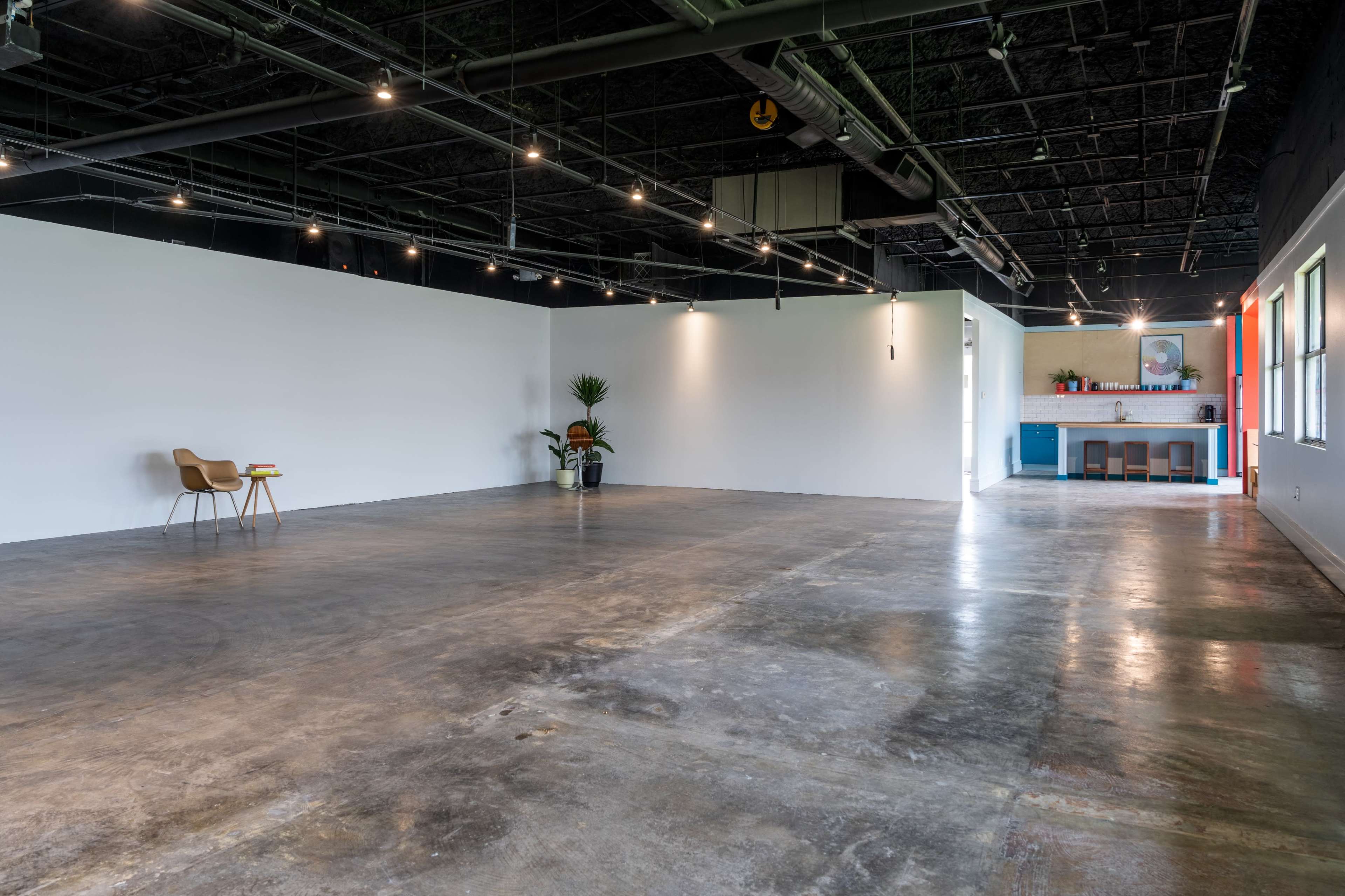 A spacious, empty room with polished concrete floors, a single chair and table in one corner, and a kitchenette area visible in the background.
