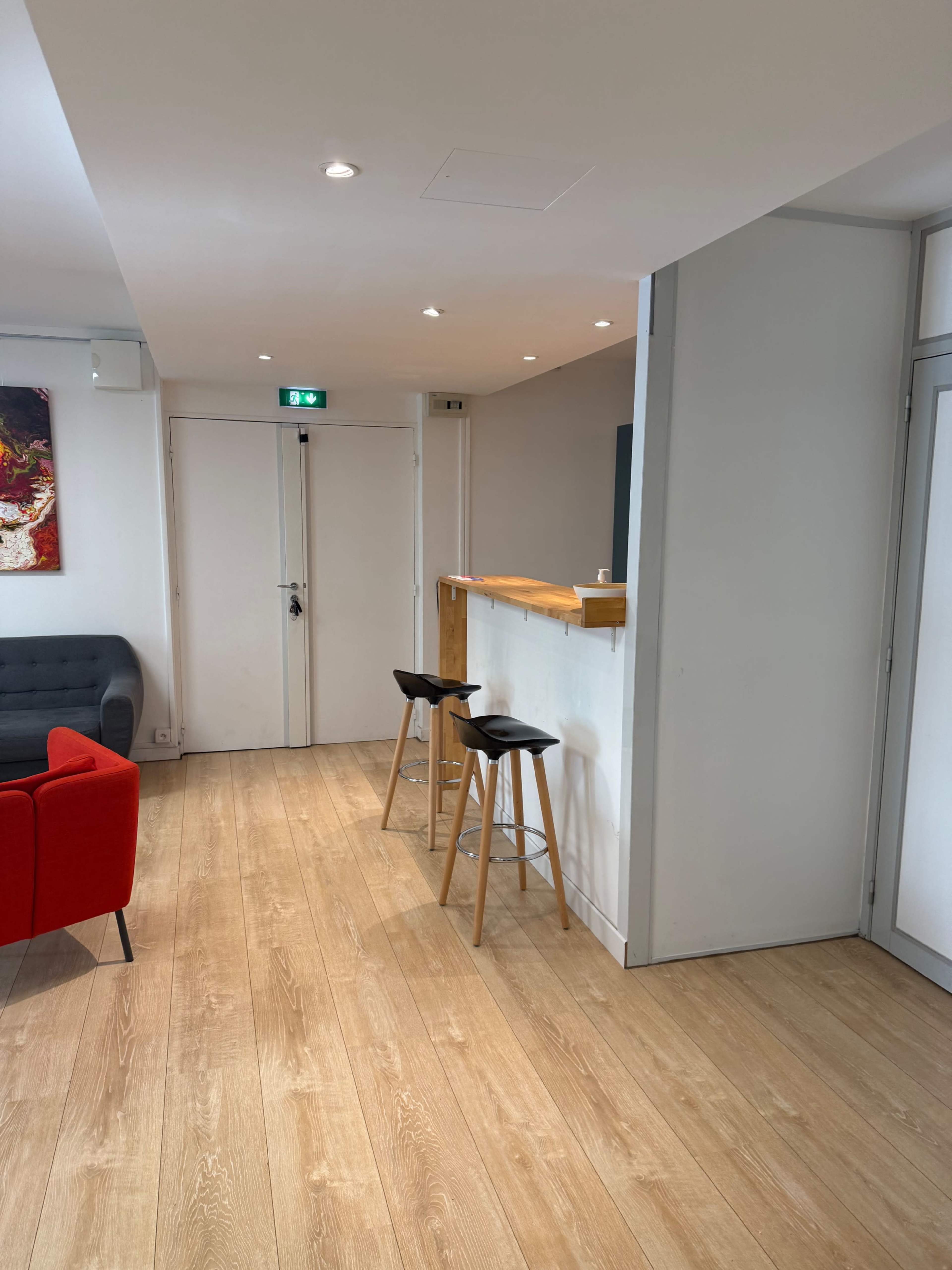 The image shows a modern interior space featuring a gray sofa, a red chair, a wooden bar counter with two stools, and light wood flooring.