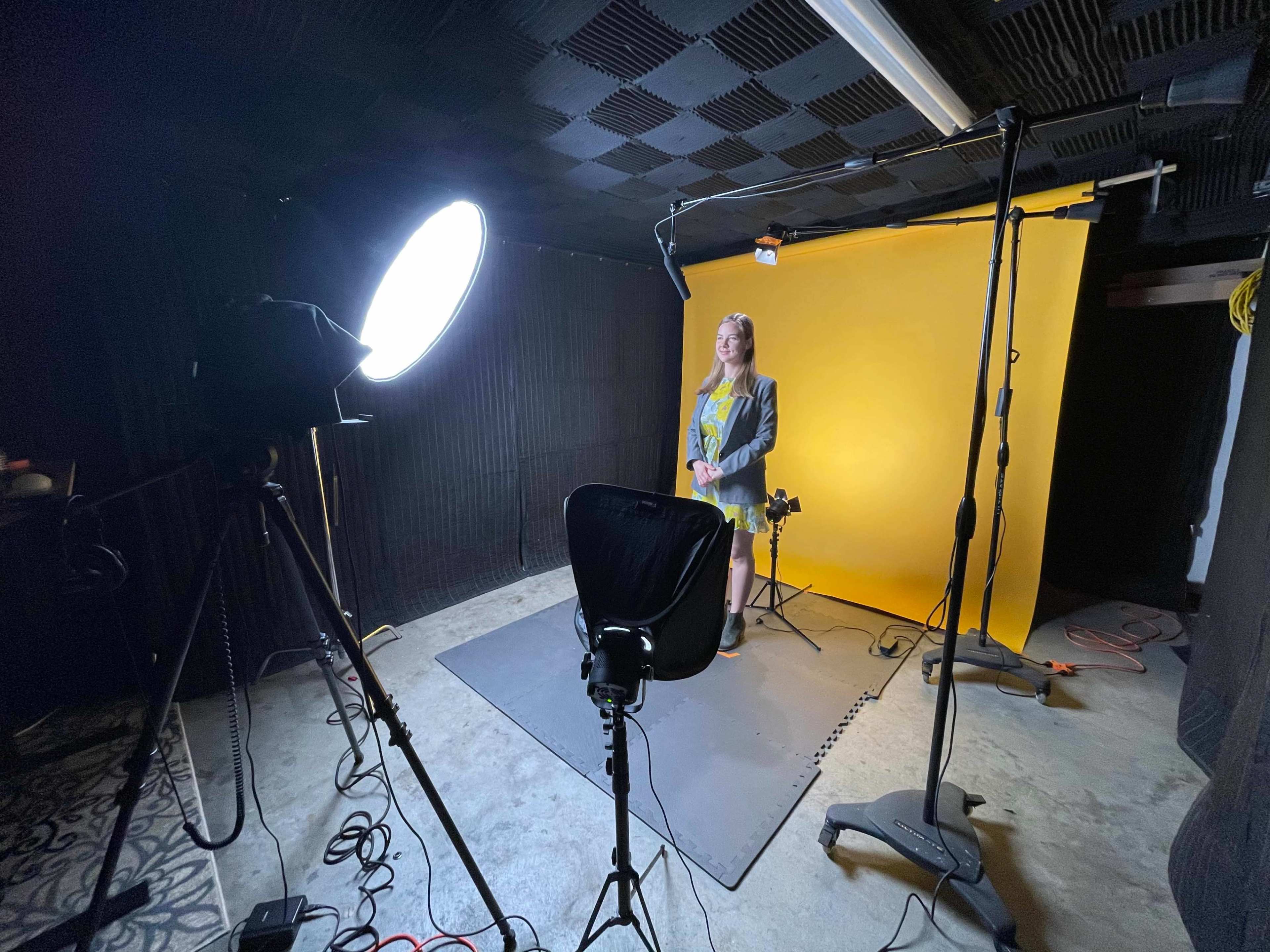 A person stands in front of a bright yellow backdrop in a photography studio, surrounded by professional lighting equipment.