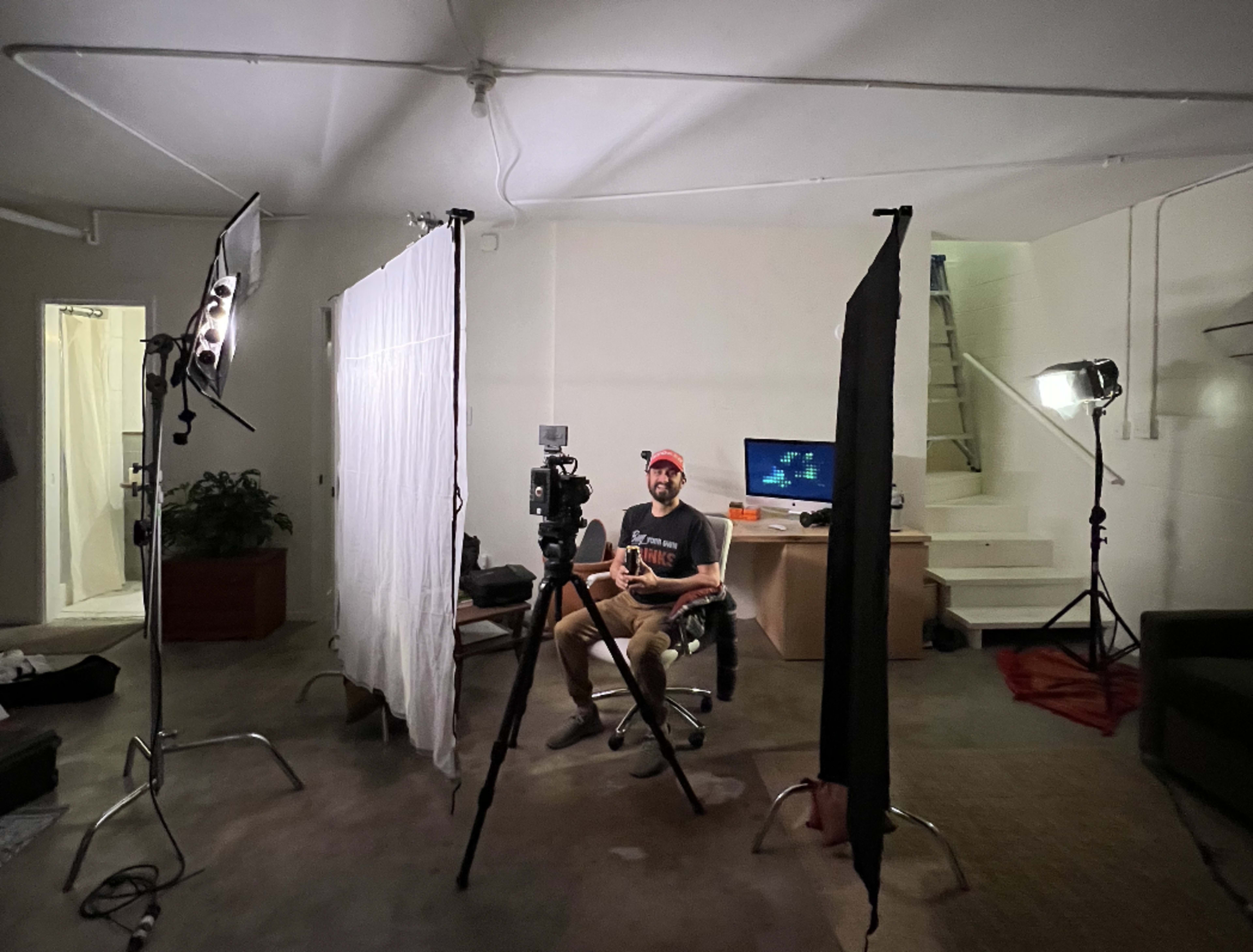 A person sits on a chair in a room set up for video recording, surrounded by lighting equipment and backdrops.