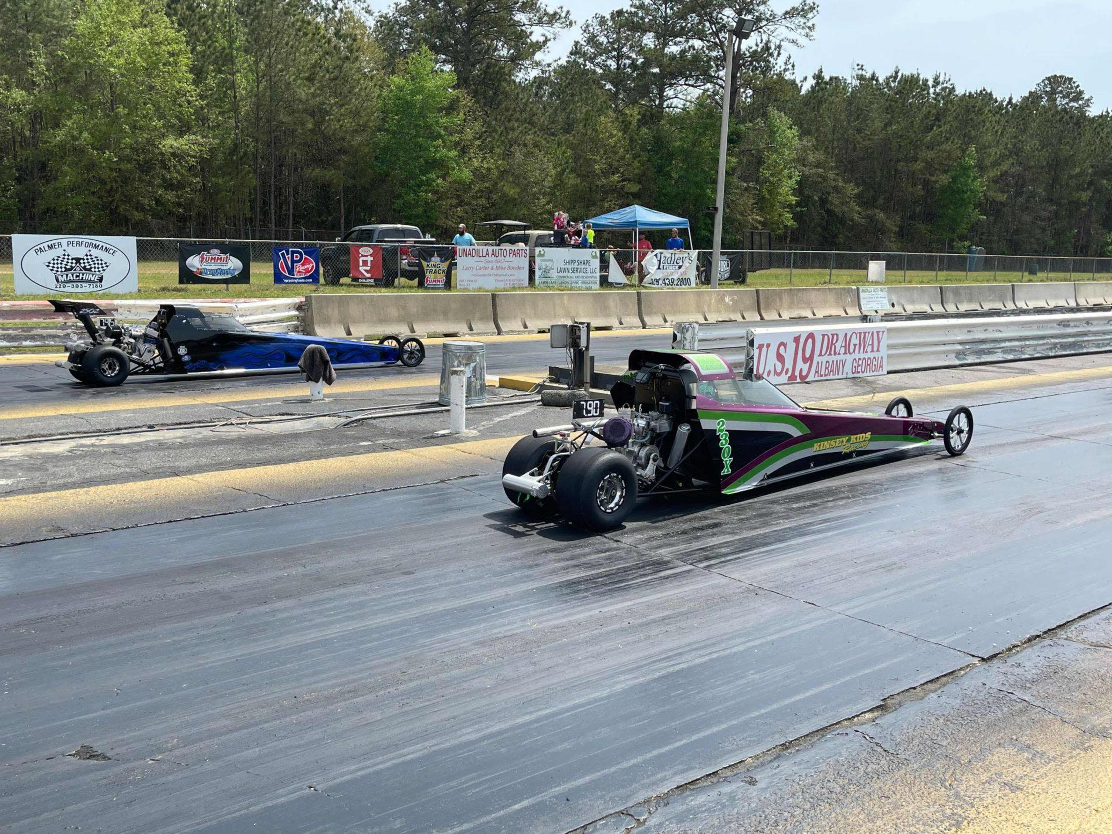 Two drag racing cars are lined up at the starting line on a racetrack, with one car in purple and green and the other in blue, while spectators watch from the sidelines under tents.