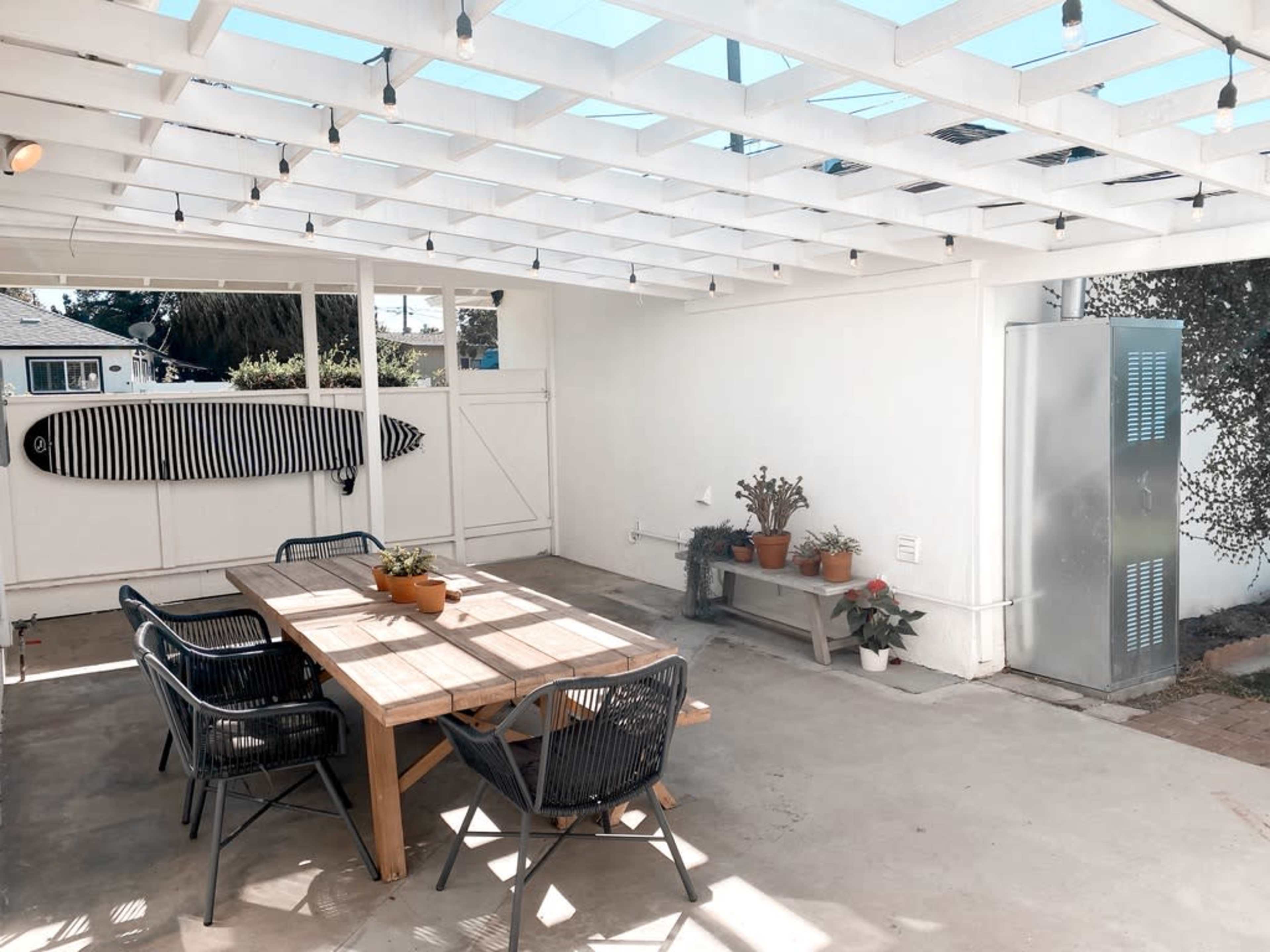A covered outdoor patio area with a wooden table, black chairs, potted plants, and a surfboard mounted on the wall.