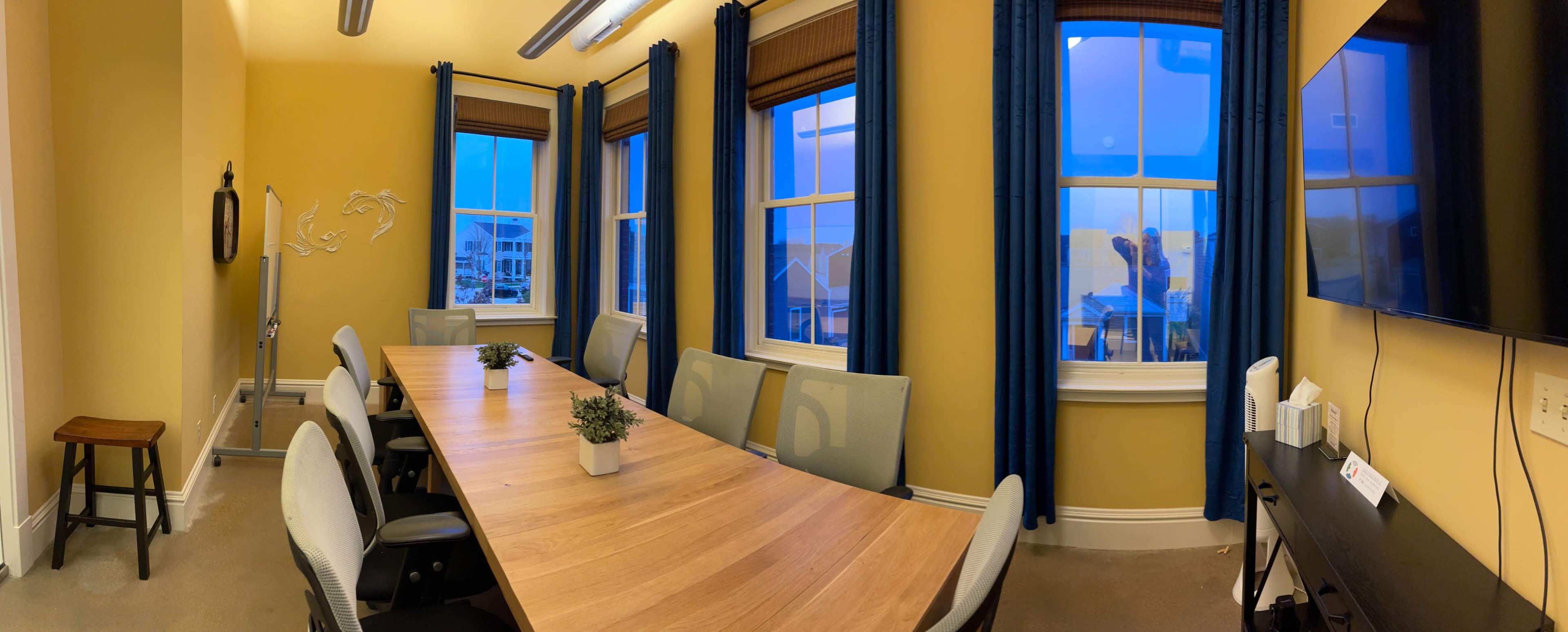 A panoramic view of a conference room features a long wooden table, several chairs, and large windows with blue curtains, illuminated by evening light.