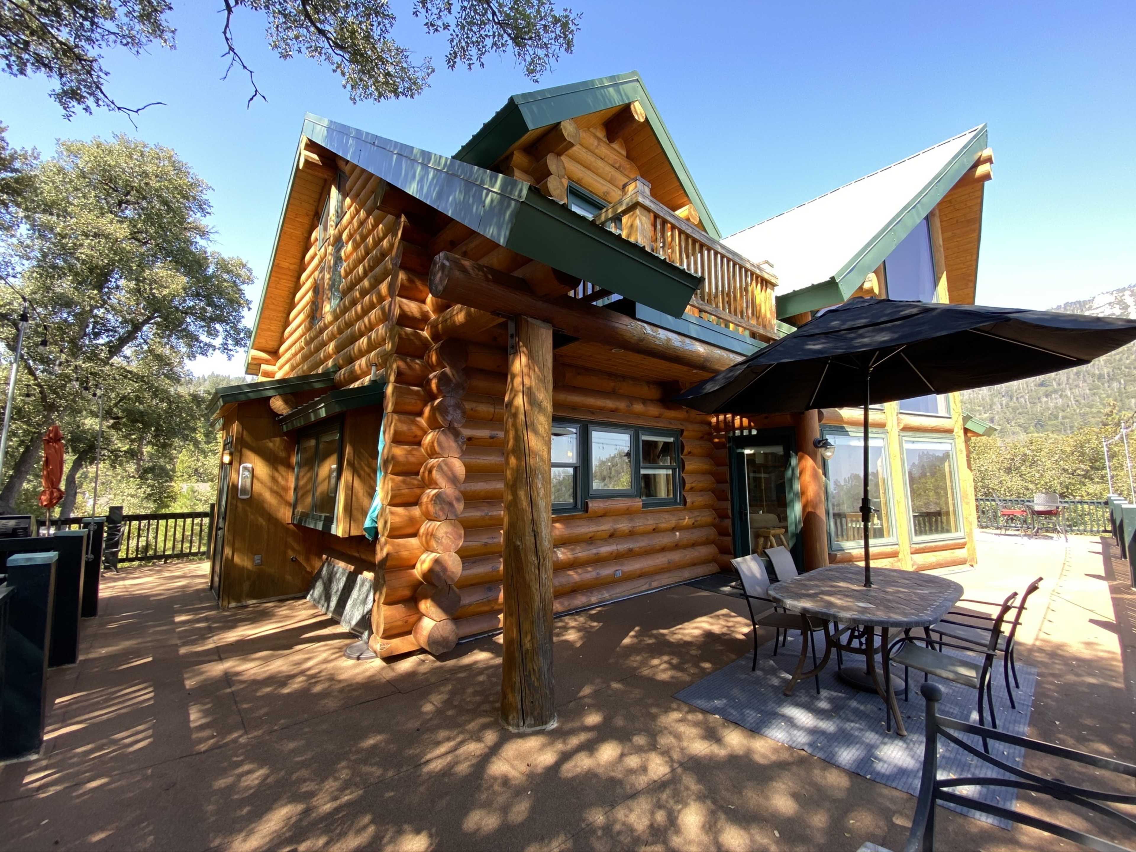 A large log cabin with a sloped roof, outdoor seating, and a view of trees and hills in the background.