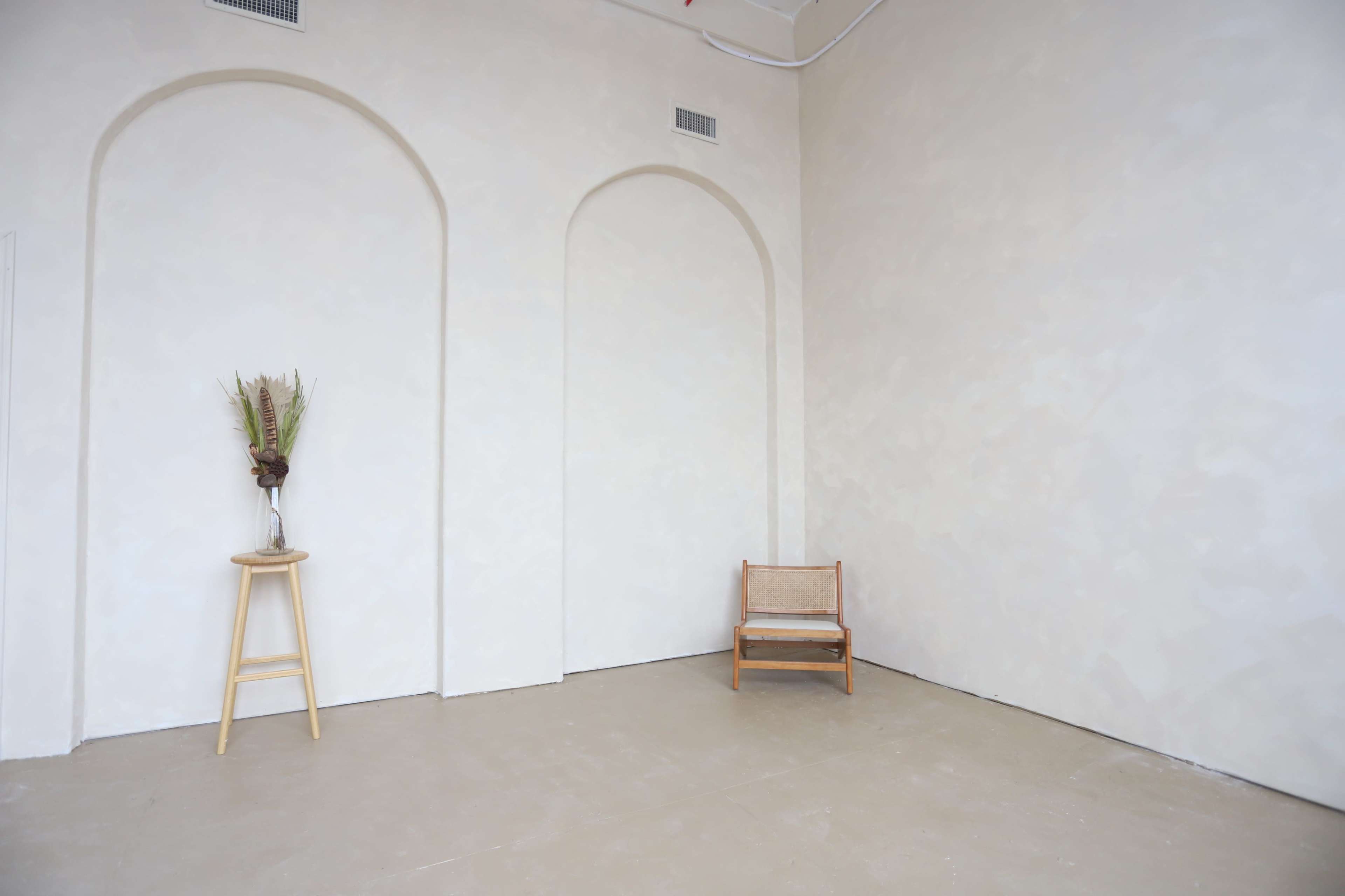 A minimalist interior with two arched wall alcoves, a small wooden chair, and a plant in a vase on a wooden stool.