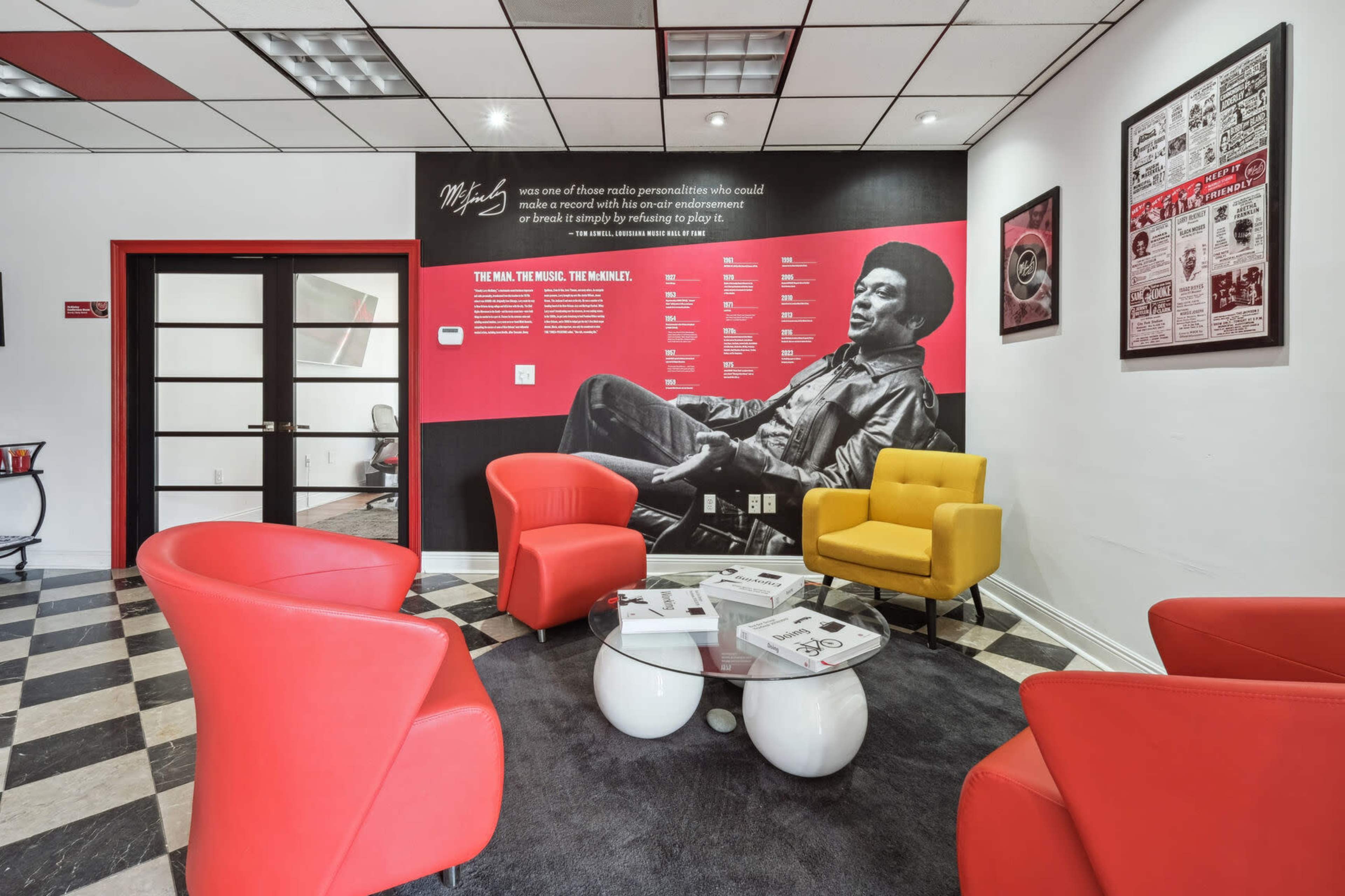 The image shows a modern lounge area with four colorful chairs and a table, alongside a wall featuring a large black-and-white mural of a musician and informational text.