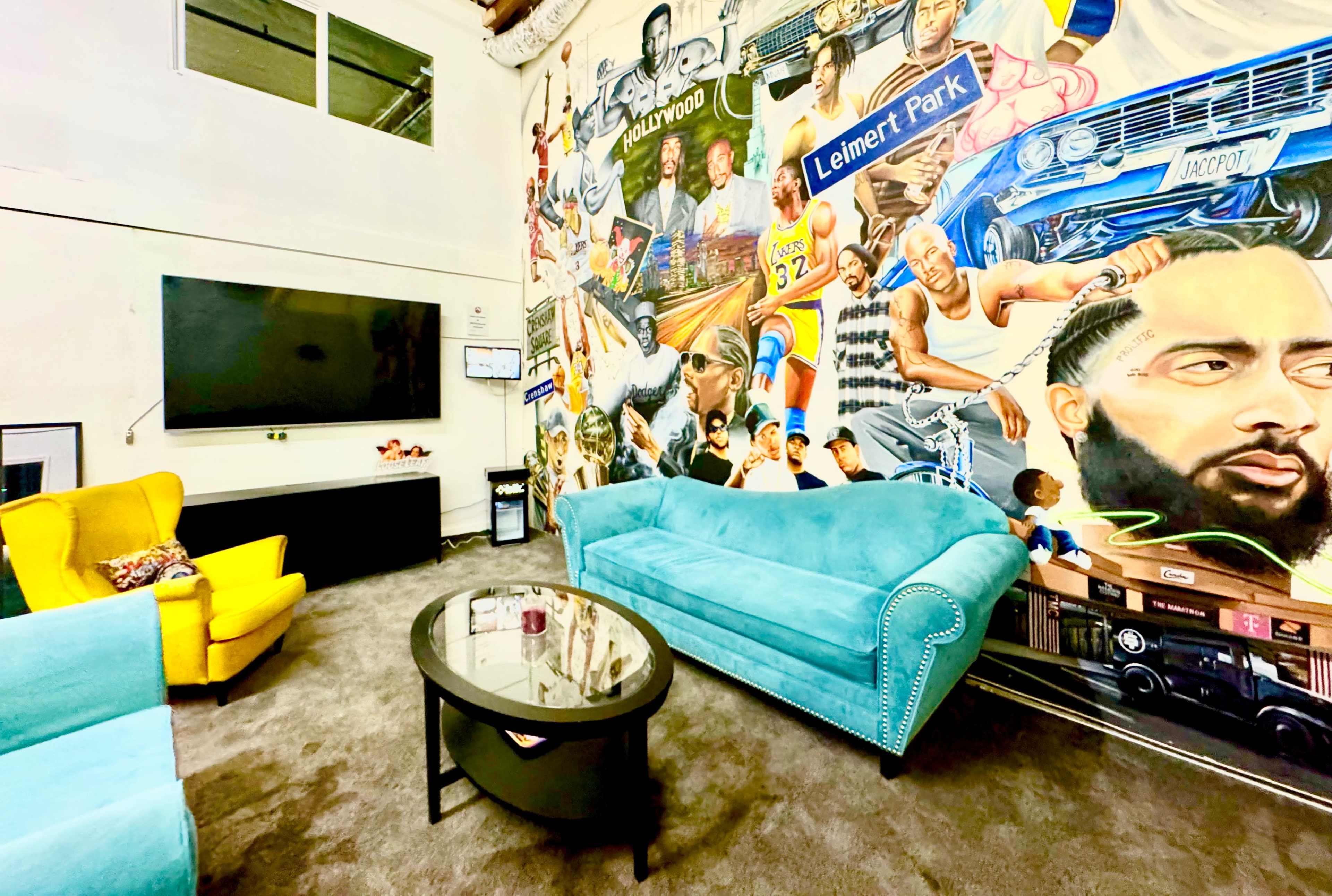 The image shows a brightly colored lounge area with turquoise and yellow chairs, a black coffee table, and a large mural depicting various cultural icons on the wall.
