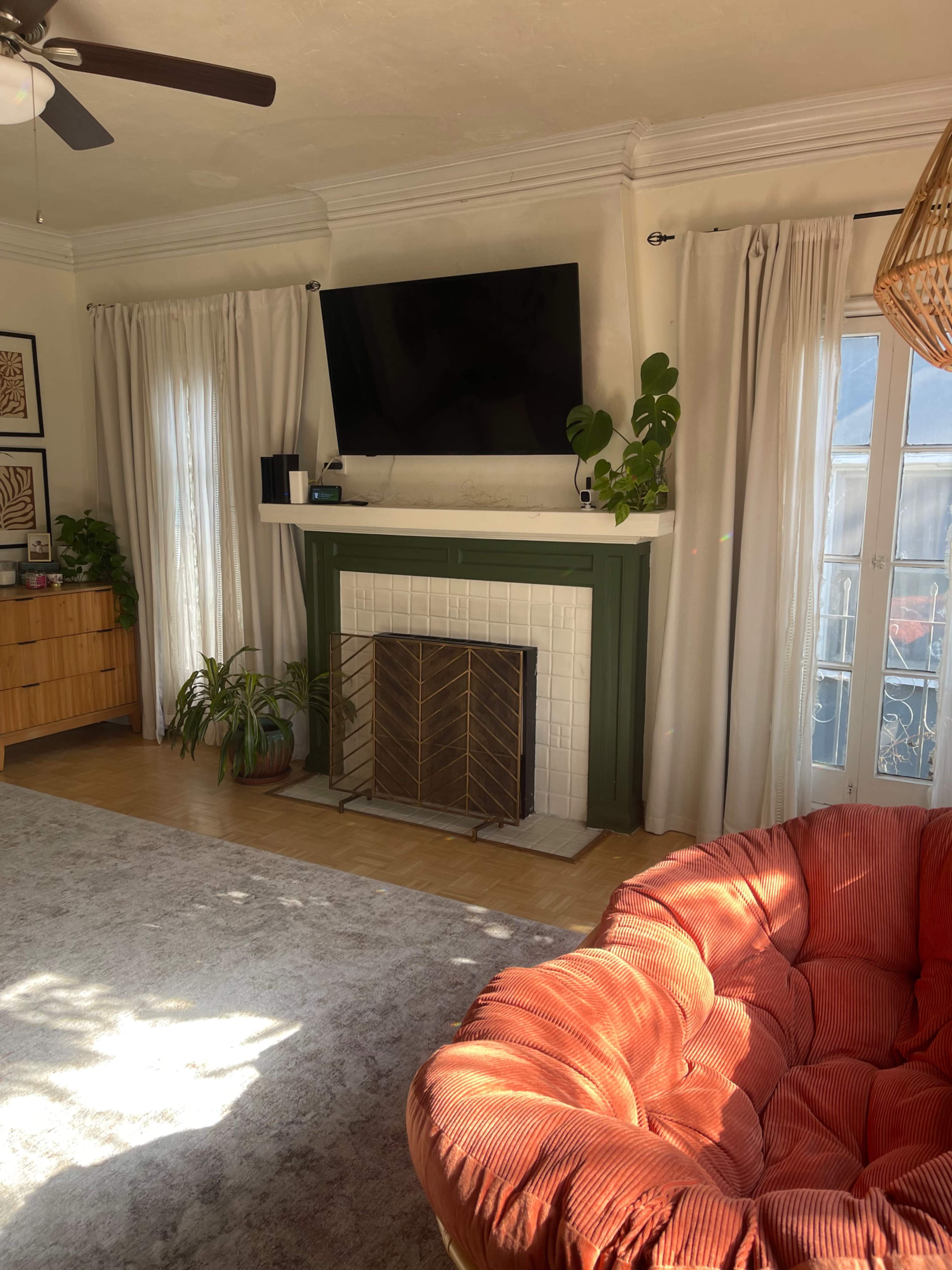 The living room features a large television mounted above a green fireplace, surrounded by white curtains, a round orange chair, and various plants.