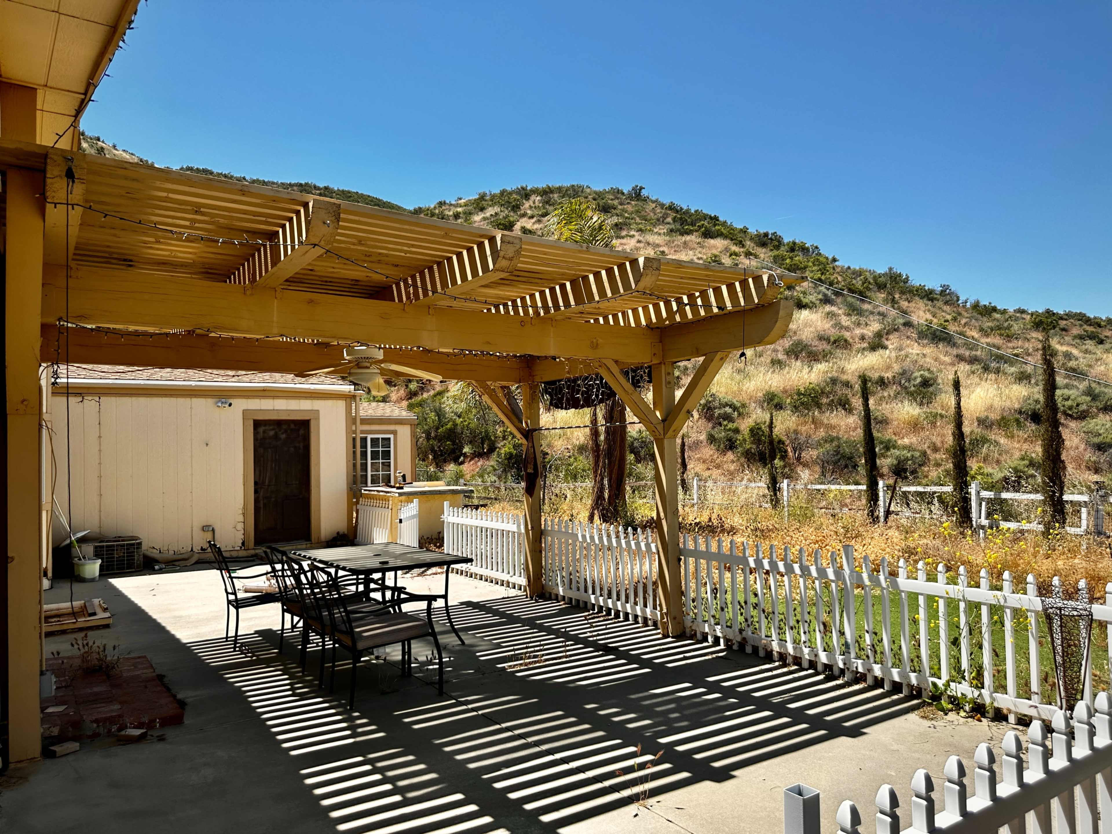 A patio with a yellow pergola and a table set beneath it is in the foreground, surrounded by a white picket fence and hills in the background.