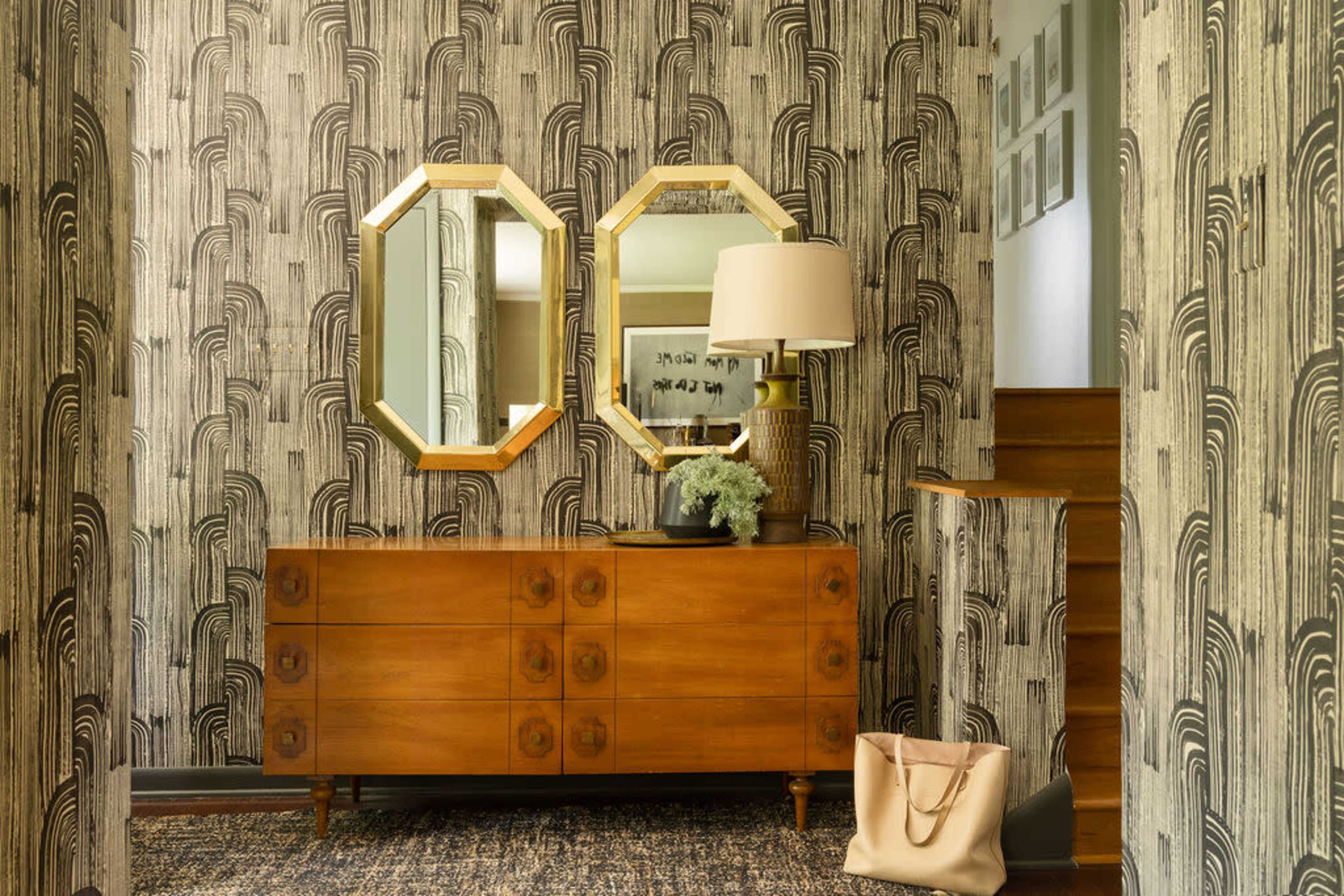 A hallway features a wooden dresser with multiple drawers, flanked by two octagonal mirrors and a lamp, against a patterned wallpaper and a staircase.