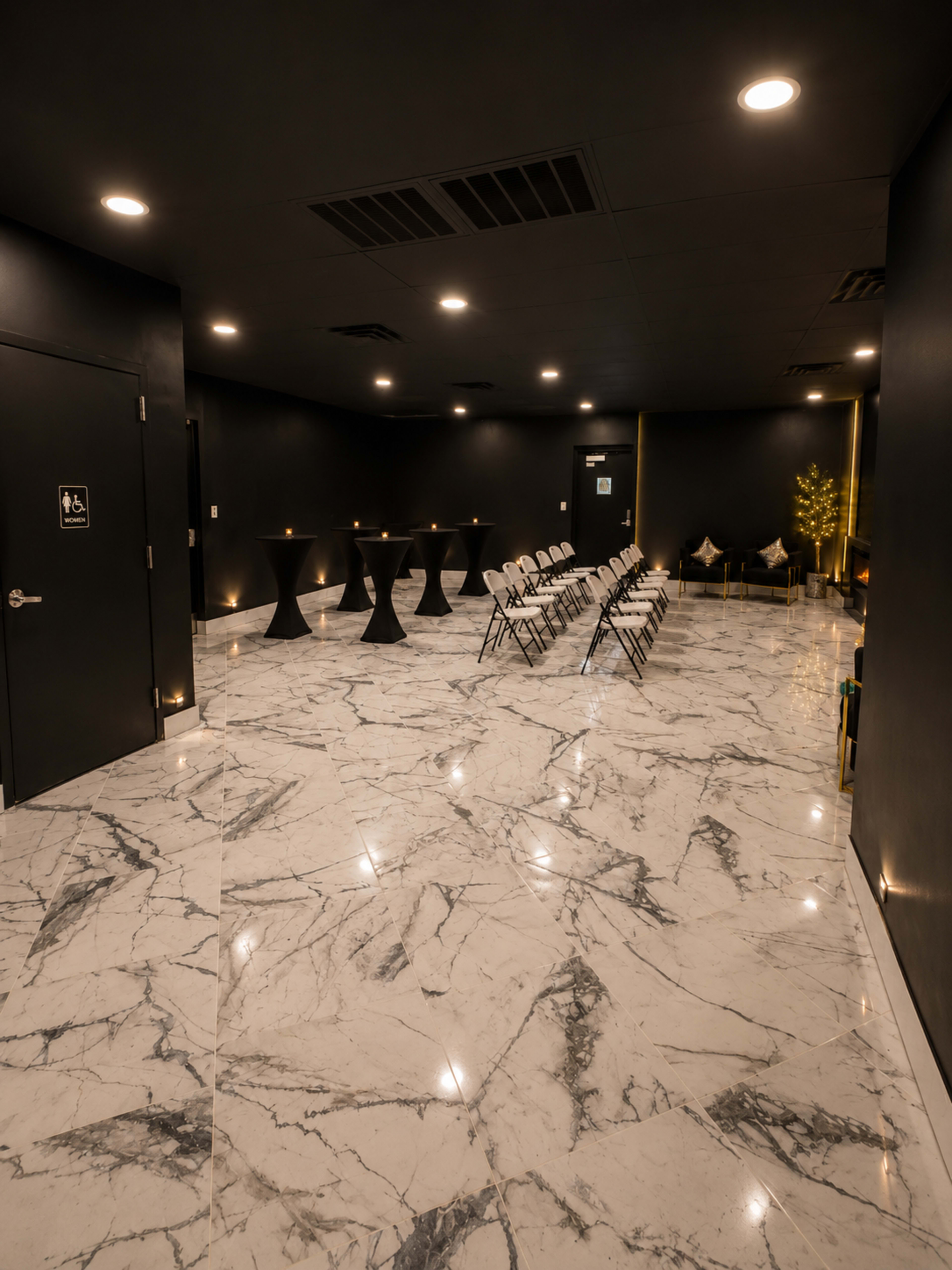 The image shows a modern event space with marble flooring, black walls, several tall black tables, and rows of white folding chairs.