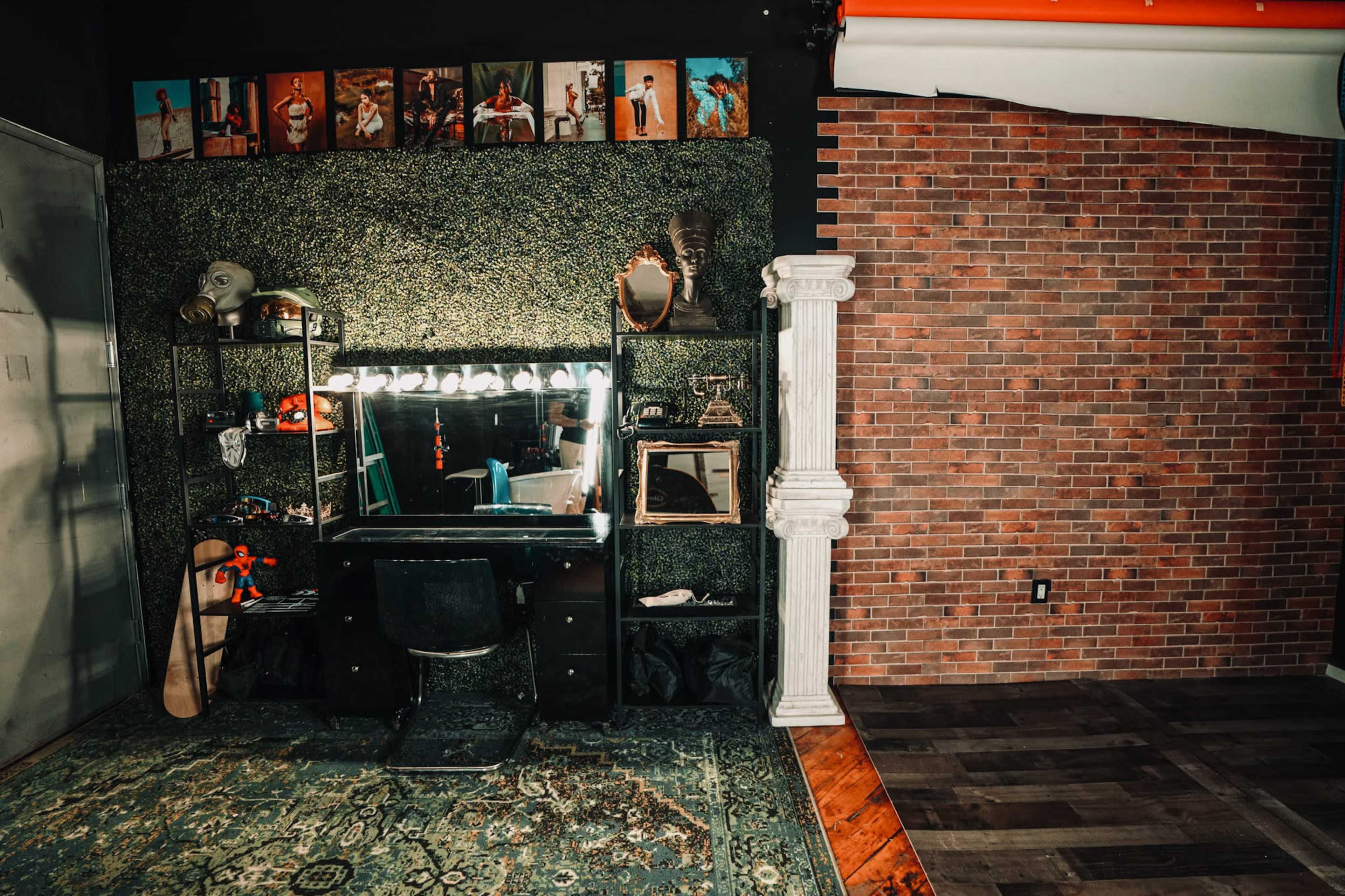 A photography studio setup featuring a dark wall with a mirror and lights, a black vanity, and a display of framed photographs above a decorative shelf.
