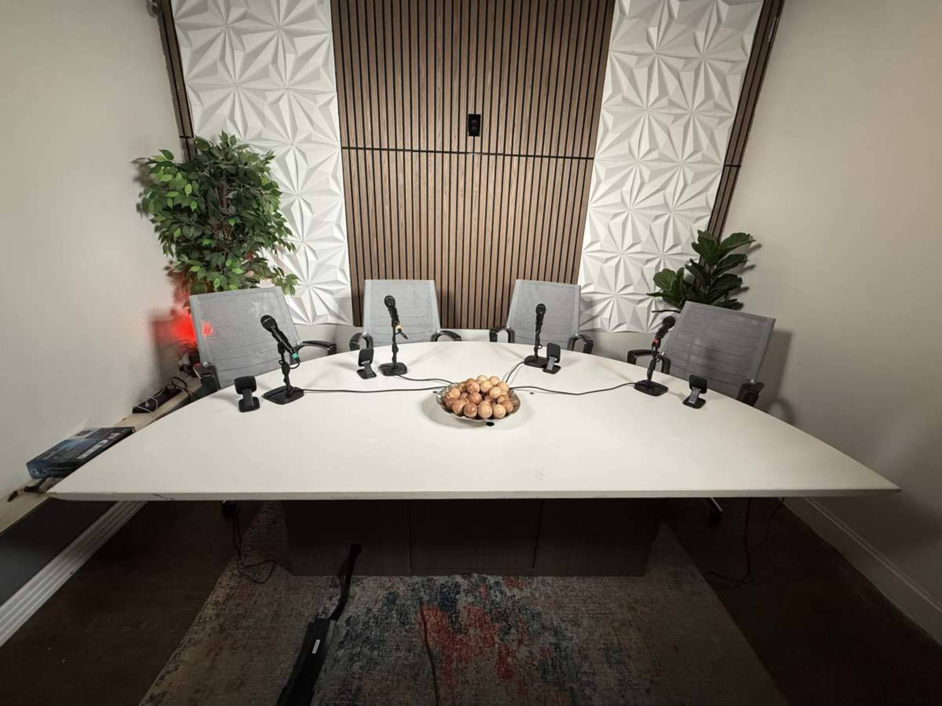 A rectangular conference table with four chairs and microphones is set up in a room featuring decorative plants and a textured wall.