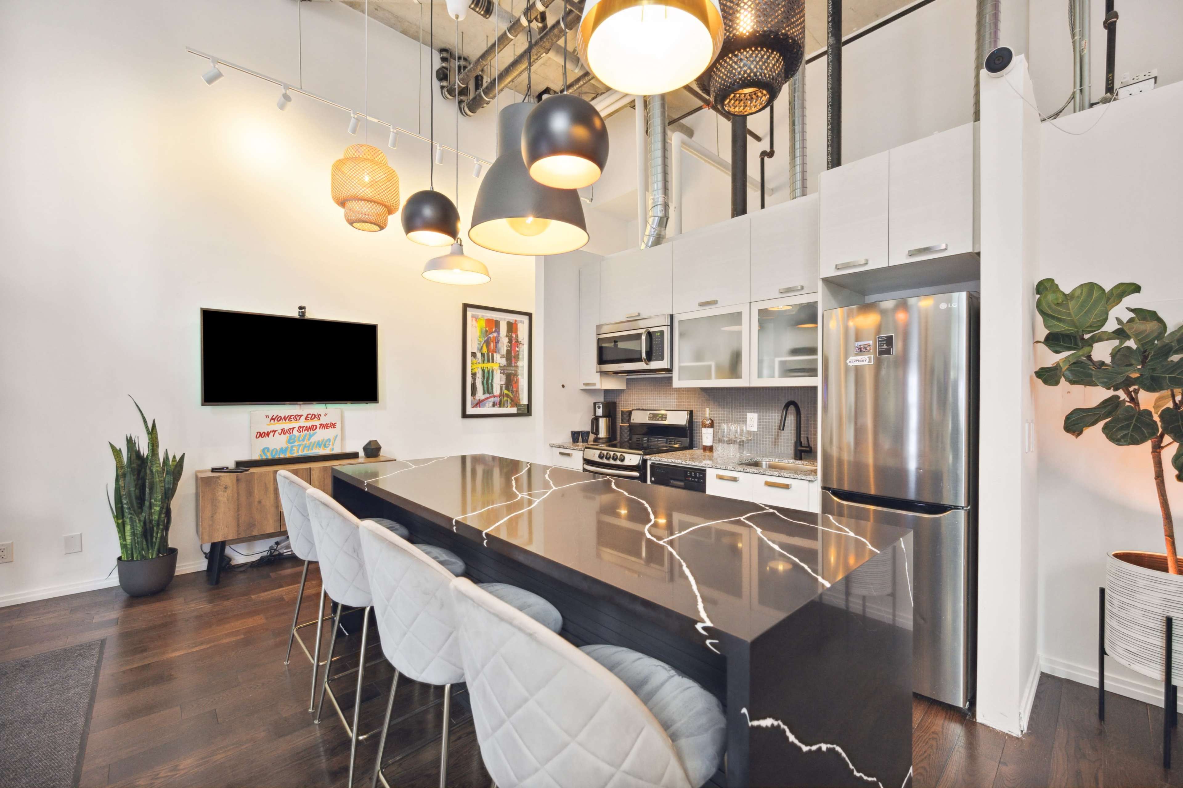 The image shows a modern kitchen with a large black countertop, several hanging light fixtures, and a wall-mounted television.