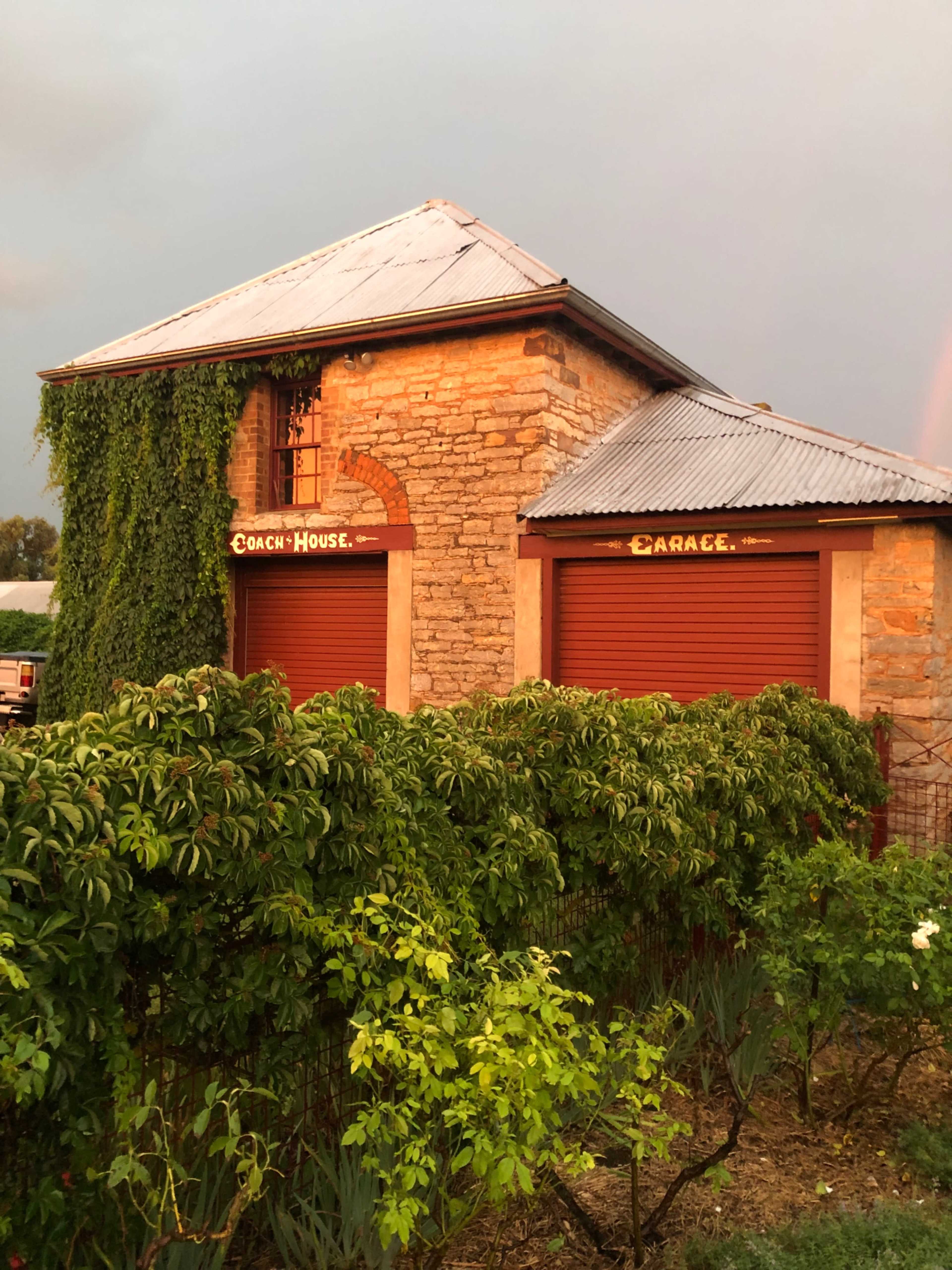A stone building with a slanted roof features red garage doors and is partially covered with greenery.