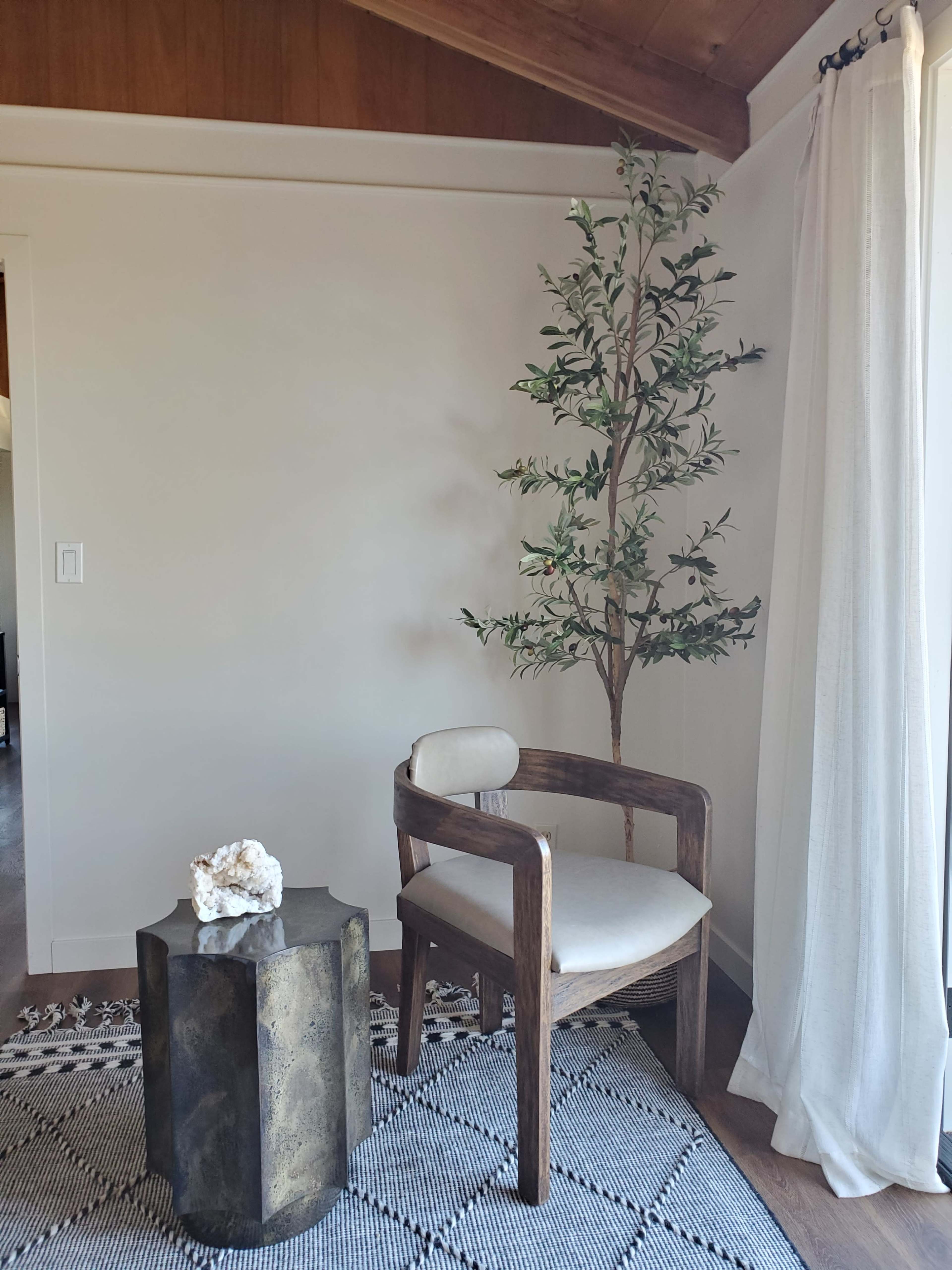 A wooden chair next to a small metal side table, with a potted plant and a light curtain in a well-lit room.