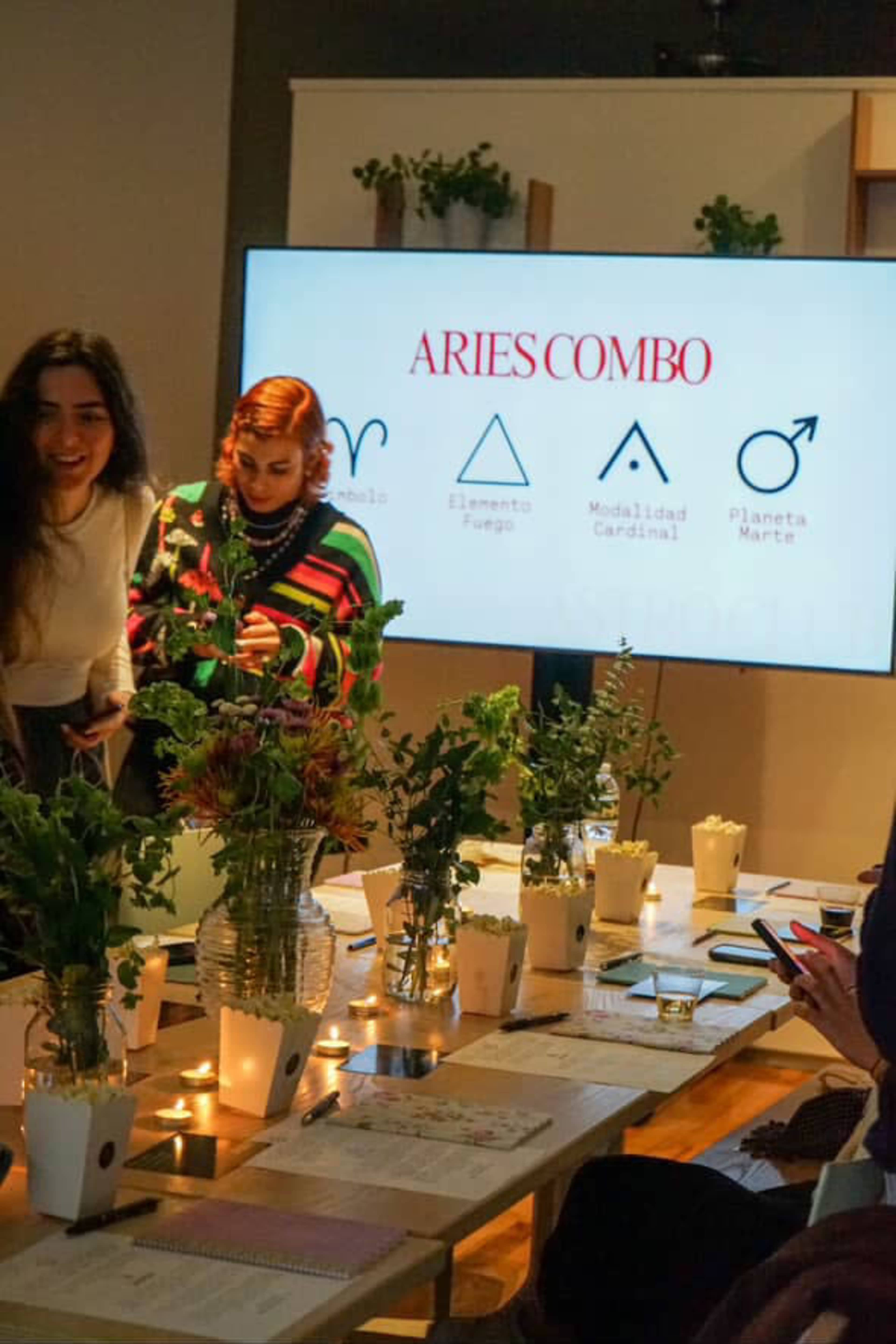 The image shows a lively gathering around a table decorated with plants and candles, with a large screen displaying information about "Aries Combo."