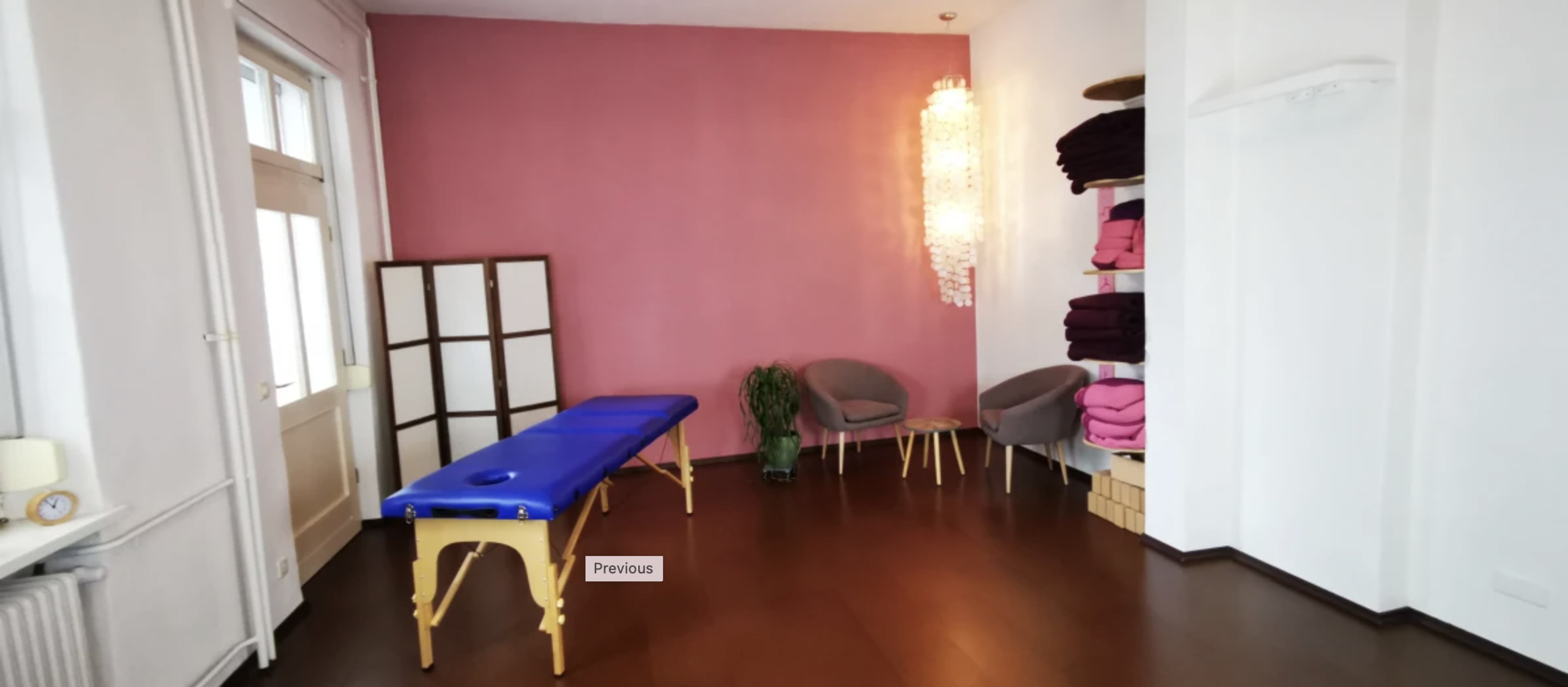 A massage therapy room featuring a blue massage table, a potted plant, two gray chairs, a shelving unit with neatly stacked towels, and a pink accent wall.