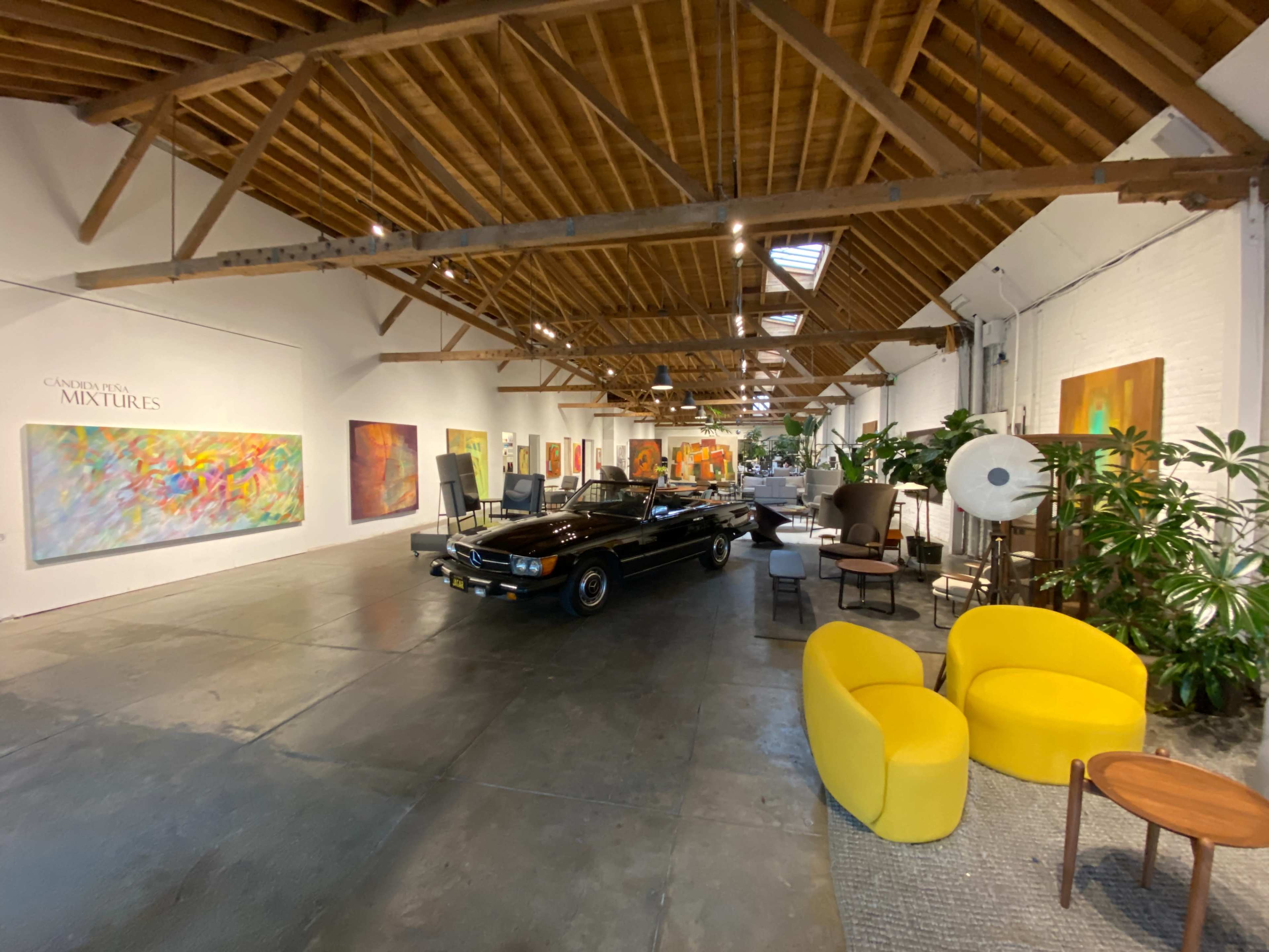 A spacious art gallery featuring colorful paintings on the walls, a classic black car in the center, and modern furniture including yellow chairs and indoor plants.