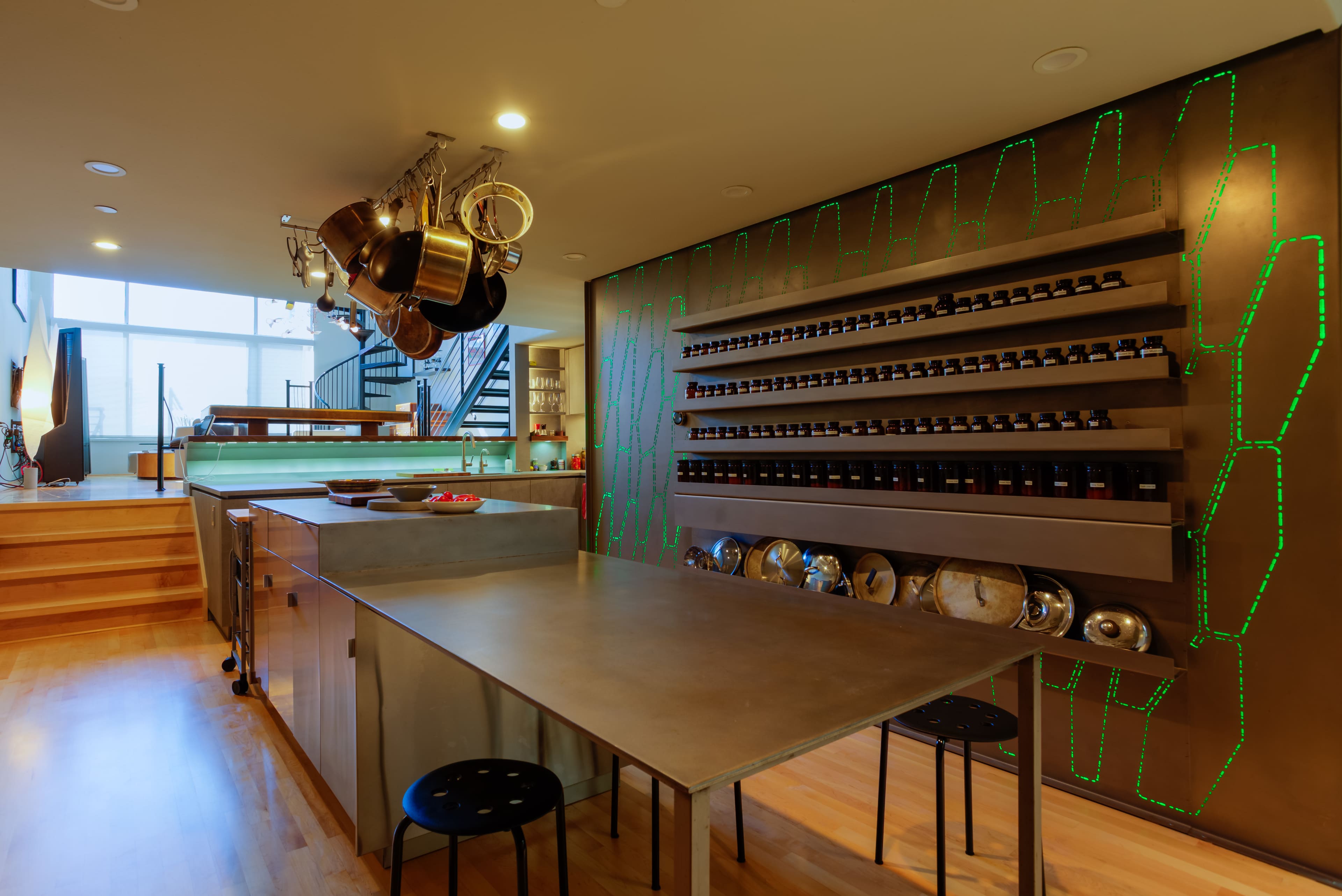 A modern kitchen with a stainless steel island, hanging pots and pans, and a wall of spice jars illuminated by green LED lights.