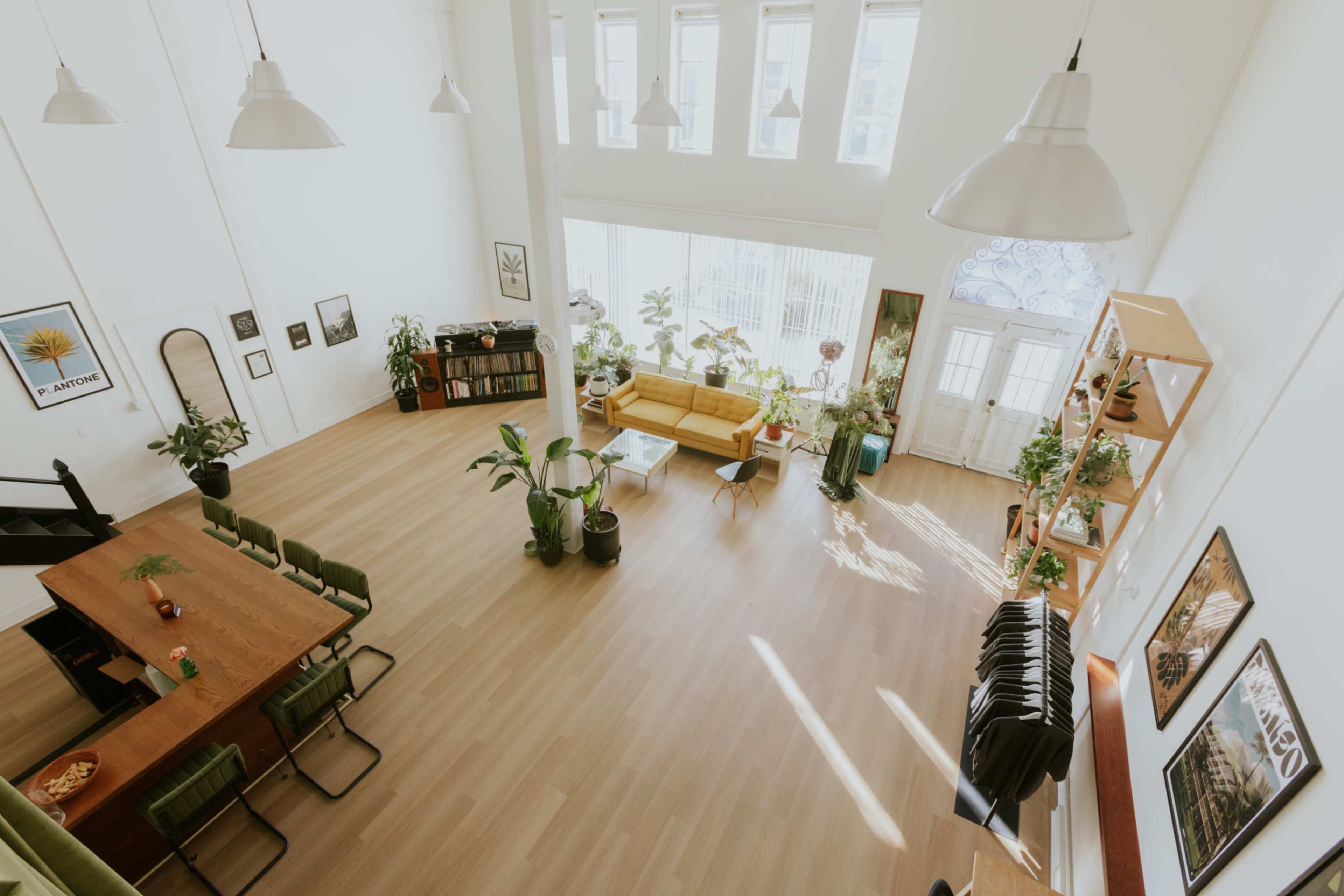 The image shows a spacious, well-lit interior with high ceilings, featuring a dining area with a long table, a living area with a yellow sofa, and various indoor plants throughout.