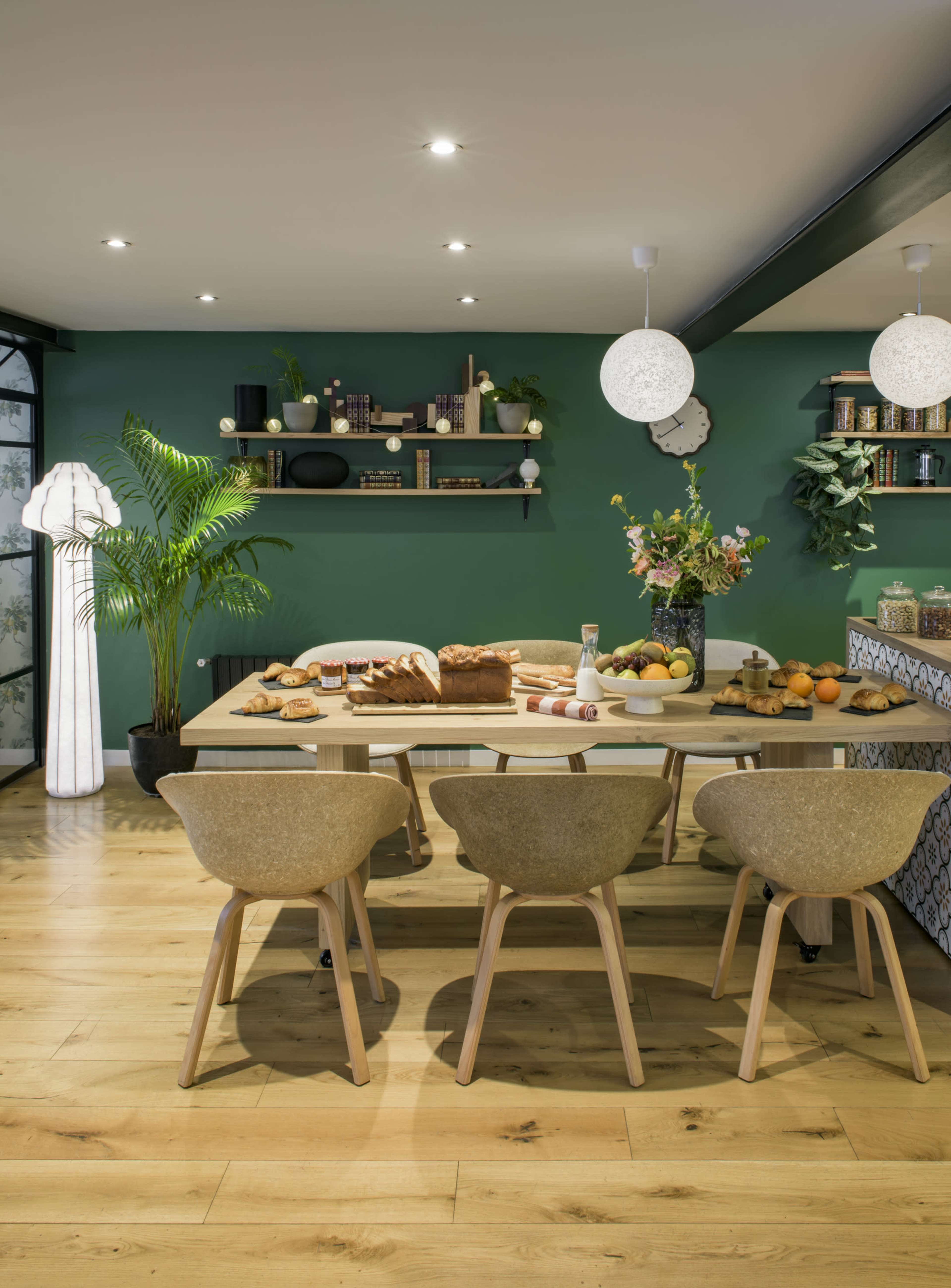 A modern dining area features a wooden table set with bread and fruit, surrounded by stylish chairs, against a green wall adorned with shelves and plants.