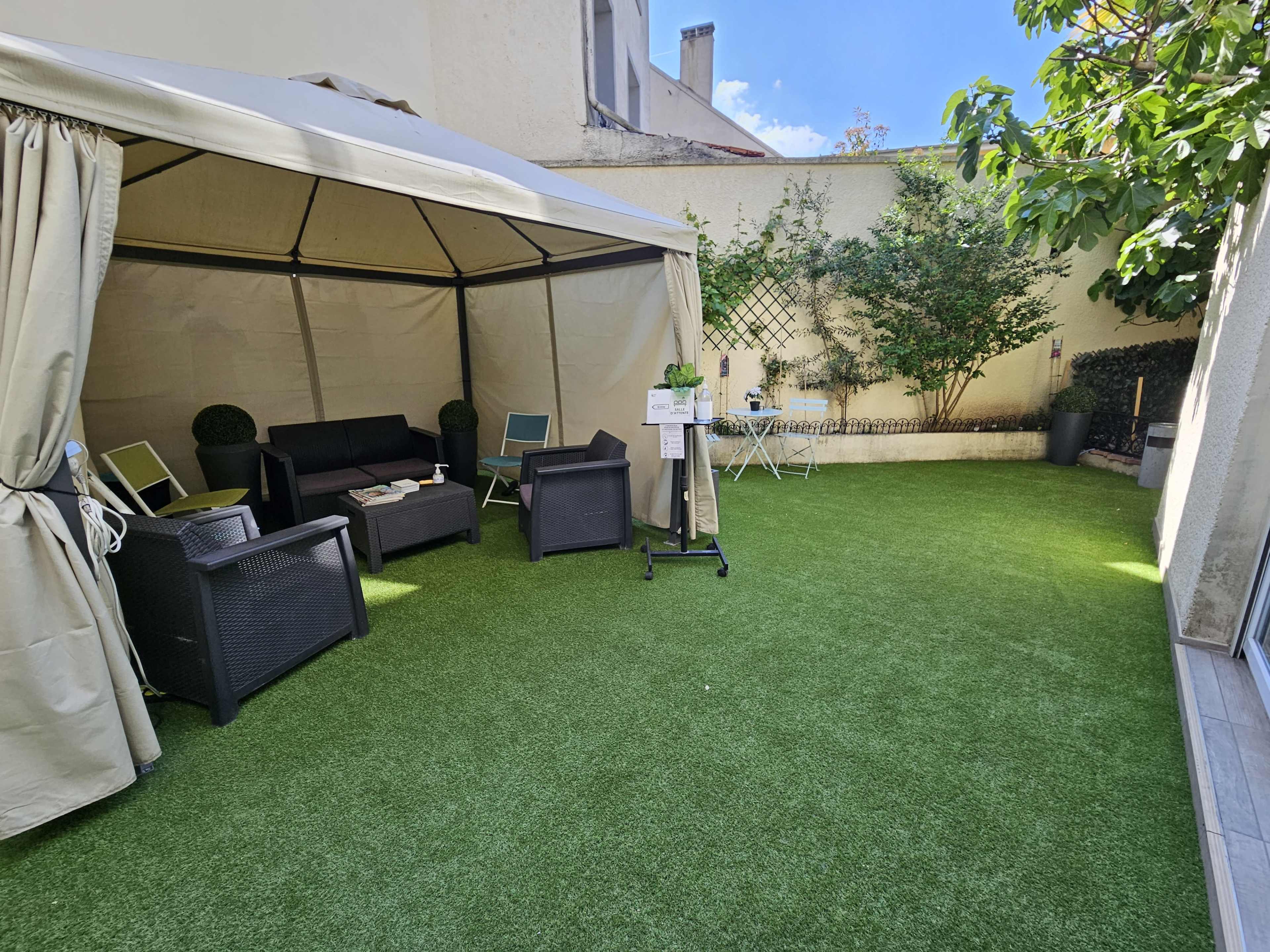 A backyard patio features a canopy tent with a seating area, artificial grass, and potted plants against a sunny backdrop.