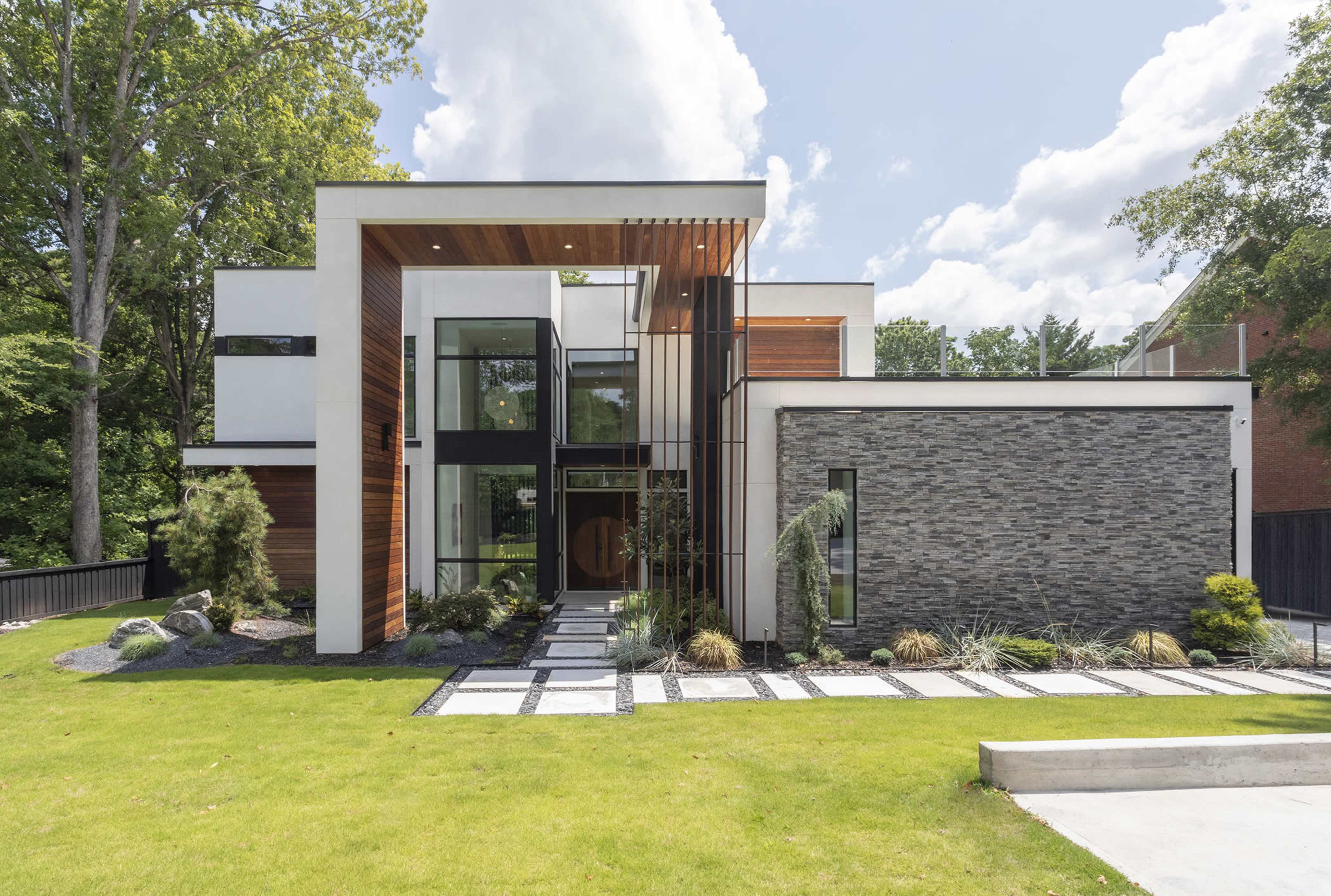 A modern house with a combination of wood and stone exteriors, featuring large windows and a symmetrical front entrance surrounded by landscaped greenery.